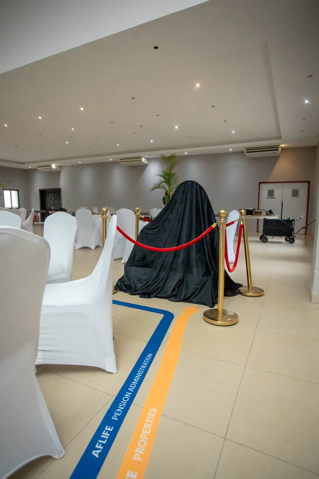 A banquet hall with white chairs, a covered object with a black cloth, and yellow and blue floor markings indicating pathways and areas for affluence life, pension administration, and properties.