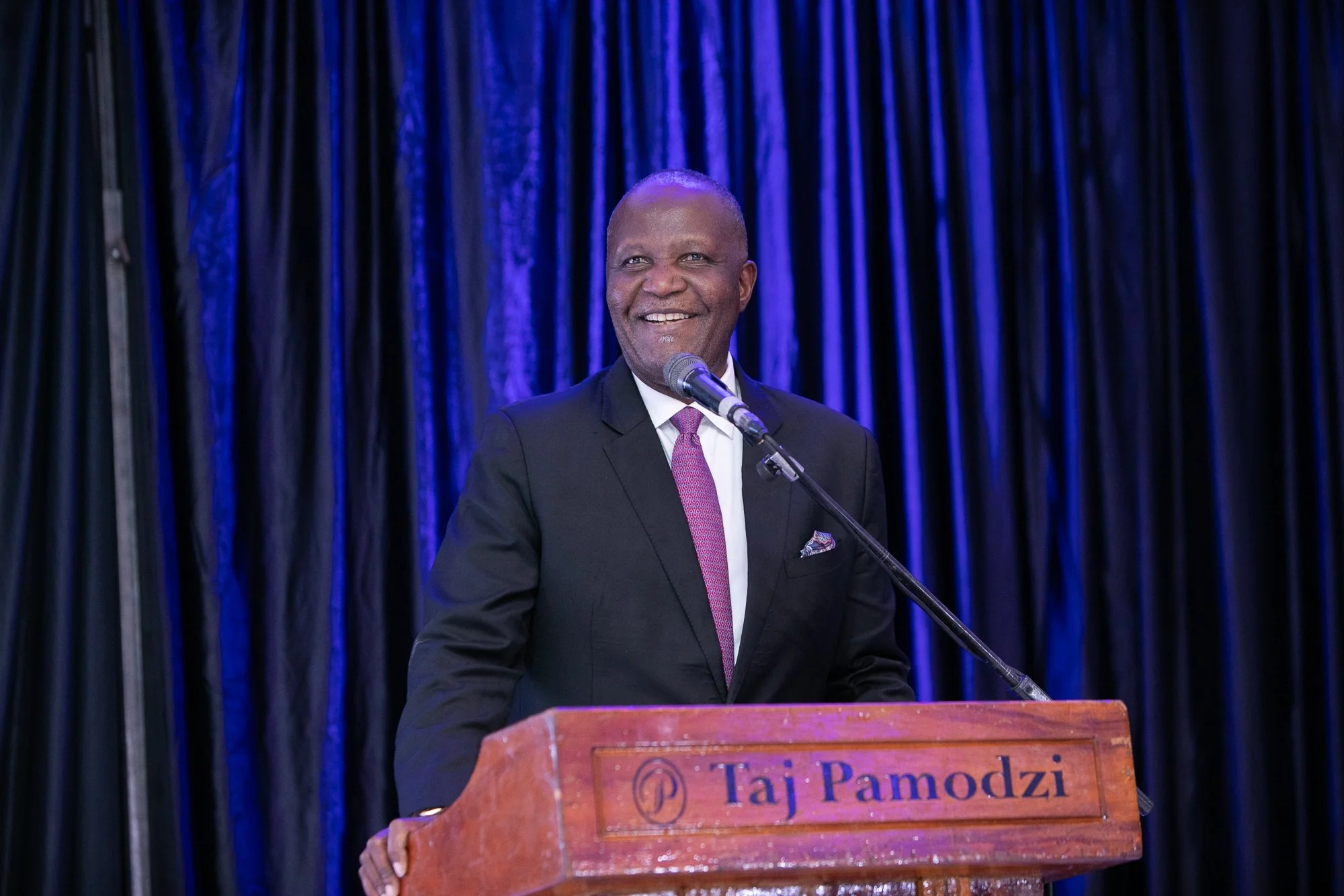 A smiling man in a black suit and pink tie speaking at a podium with an orchid logo and the words "Taj Pamodzi" in front of a dark blue curtain.