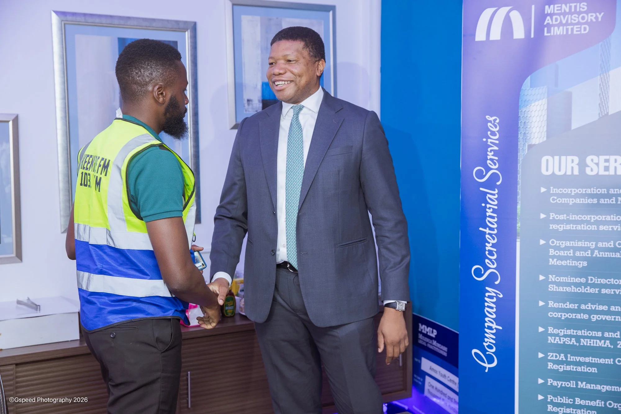 Two men, one in a suit and the other in a high-visibility vest, are shaking hands and smiling at each other in an indoor setting. Behind them is a large blue informational display and a wall with framed pictures.