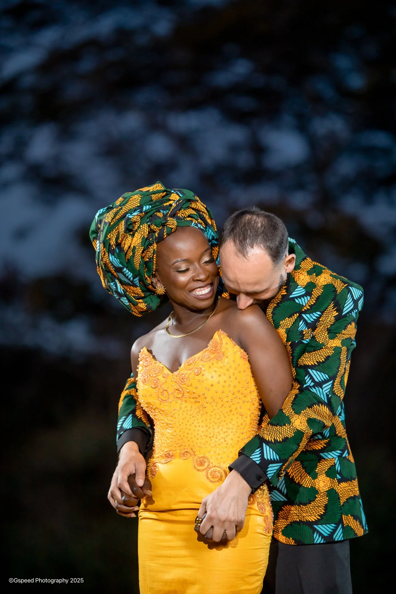 A couple dressed in bright, colorful clothing embracing each other and smiling, with a dark, blurred background.