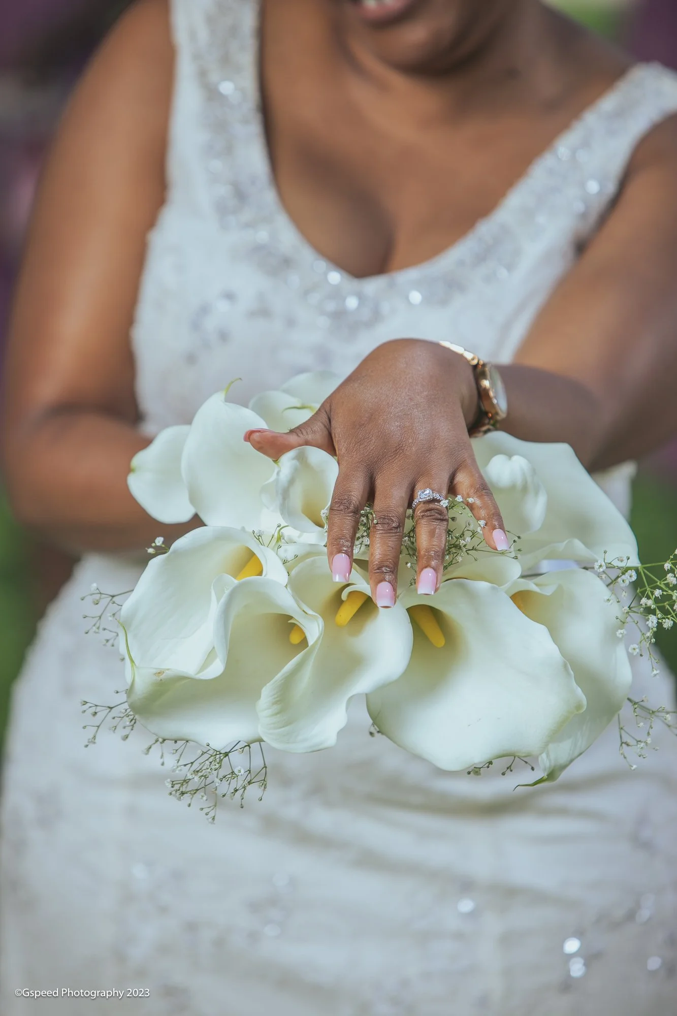 A woman in a white dress with beaded straps is holding a bouquet of white calla lilies and small white flowers. She is wearing jewelry, including a watch and rings.
