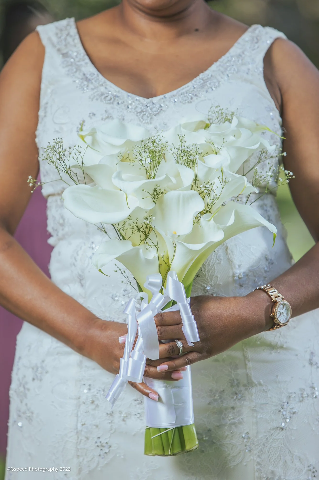 Close-up of a woman in a white lace wedding dress holding a bouquet of white calla lilies and baby's breath, with a ring on her finger and a watch on her wrist.