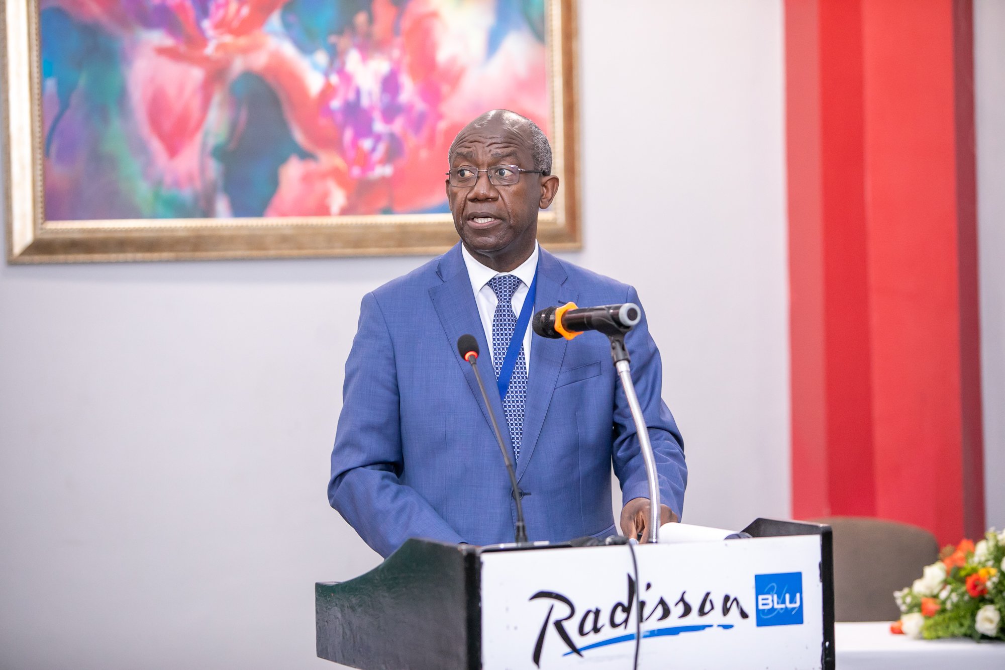 A man in a blue suit giving a speech at a conference, standing behind a podium with a sign that says "Radisson" and "BLU." A colorful abstract painting is on the wall behind him.