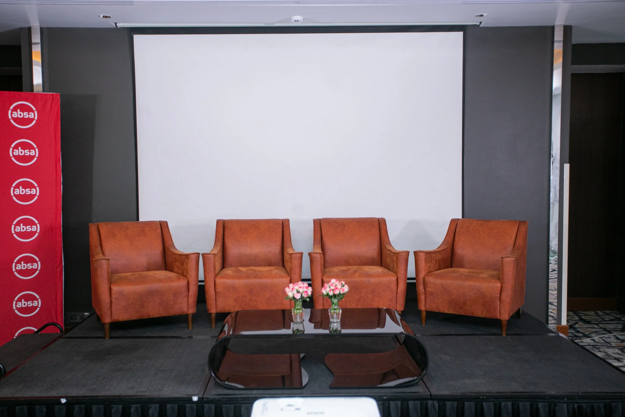 A stage setup with four brown leather armchairs, a small glass-top table with two flower vases filled with pink flowers, in front of a large white projection screen. To the left, there is a red banner with multiple white ABSA logos. The setting appea