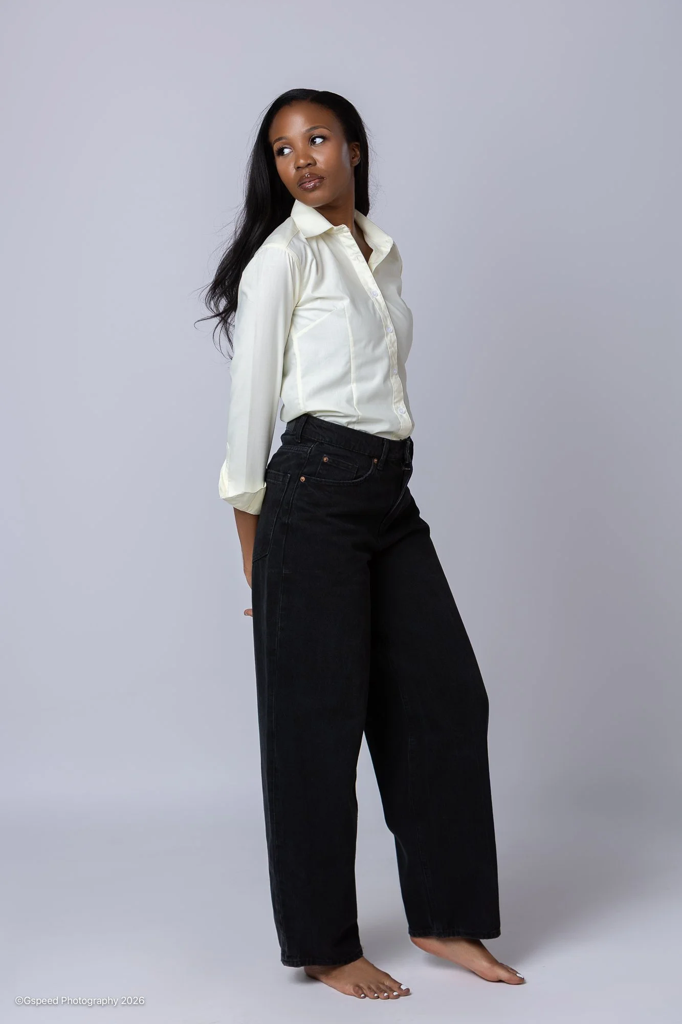 A woman with long black hair posing in front of a plain gray background, wearing a white button-up shirt and black wide-leg pants, barefoot.