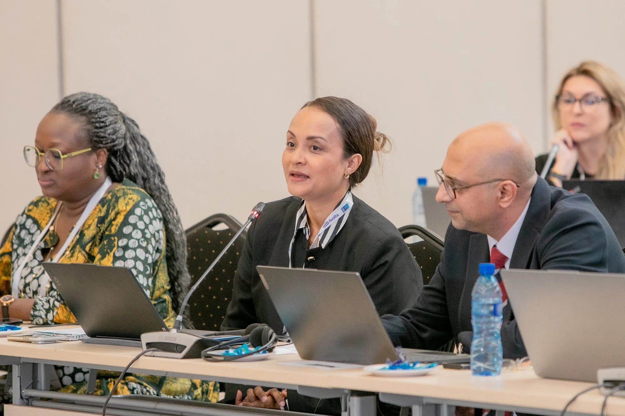 Four diverse professionals sitting at a conference table with laptops, microphones, and water bottles, engaged in a meeting or discussion.