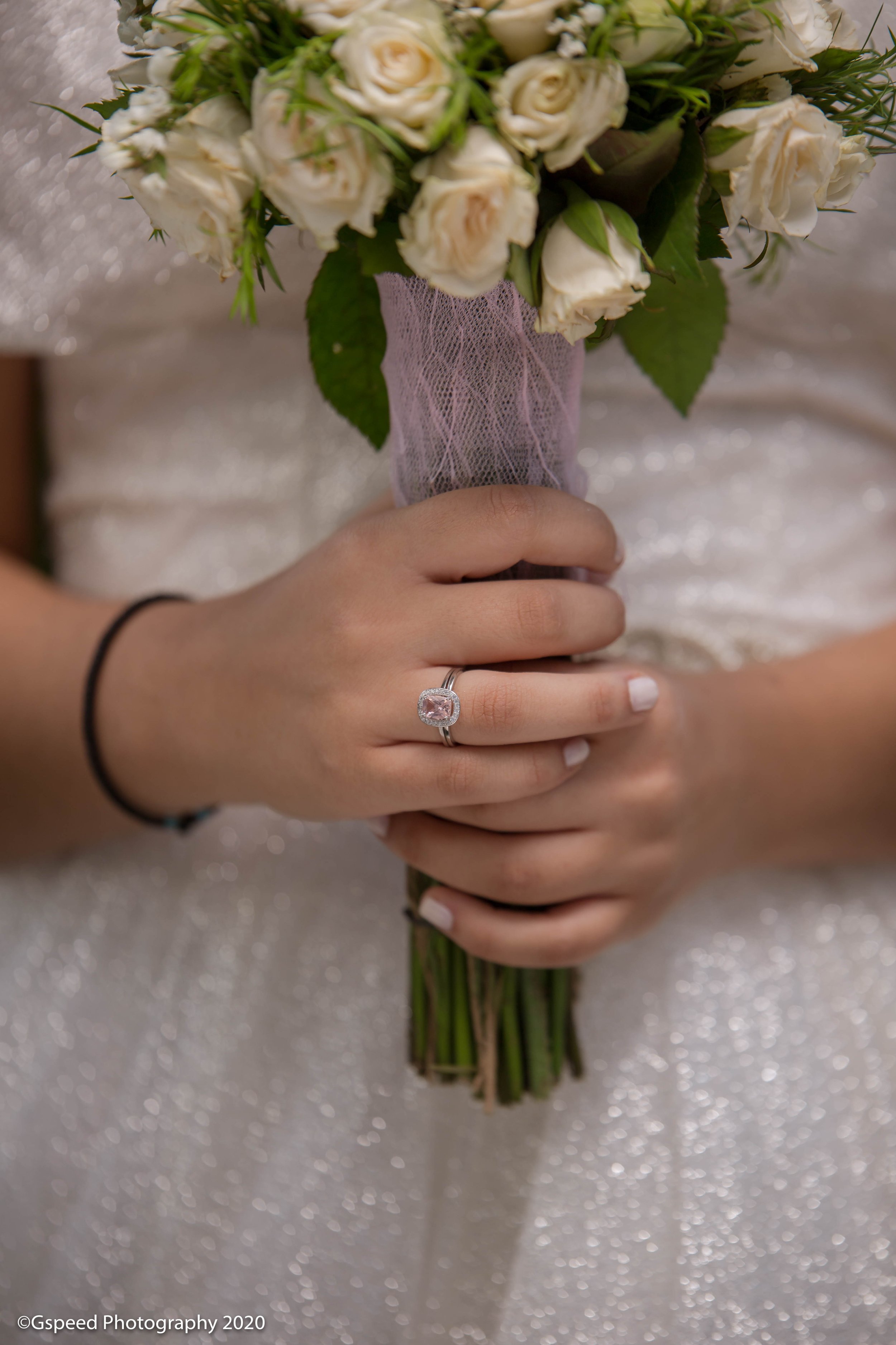 Person holding a bouquet of white roses with green leaves, wrapped in pink netting, with a silver ring on their finger.