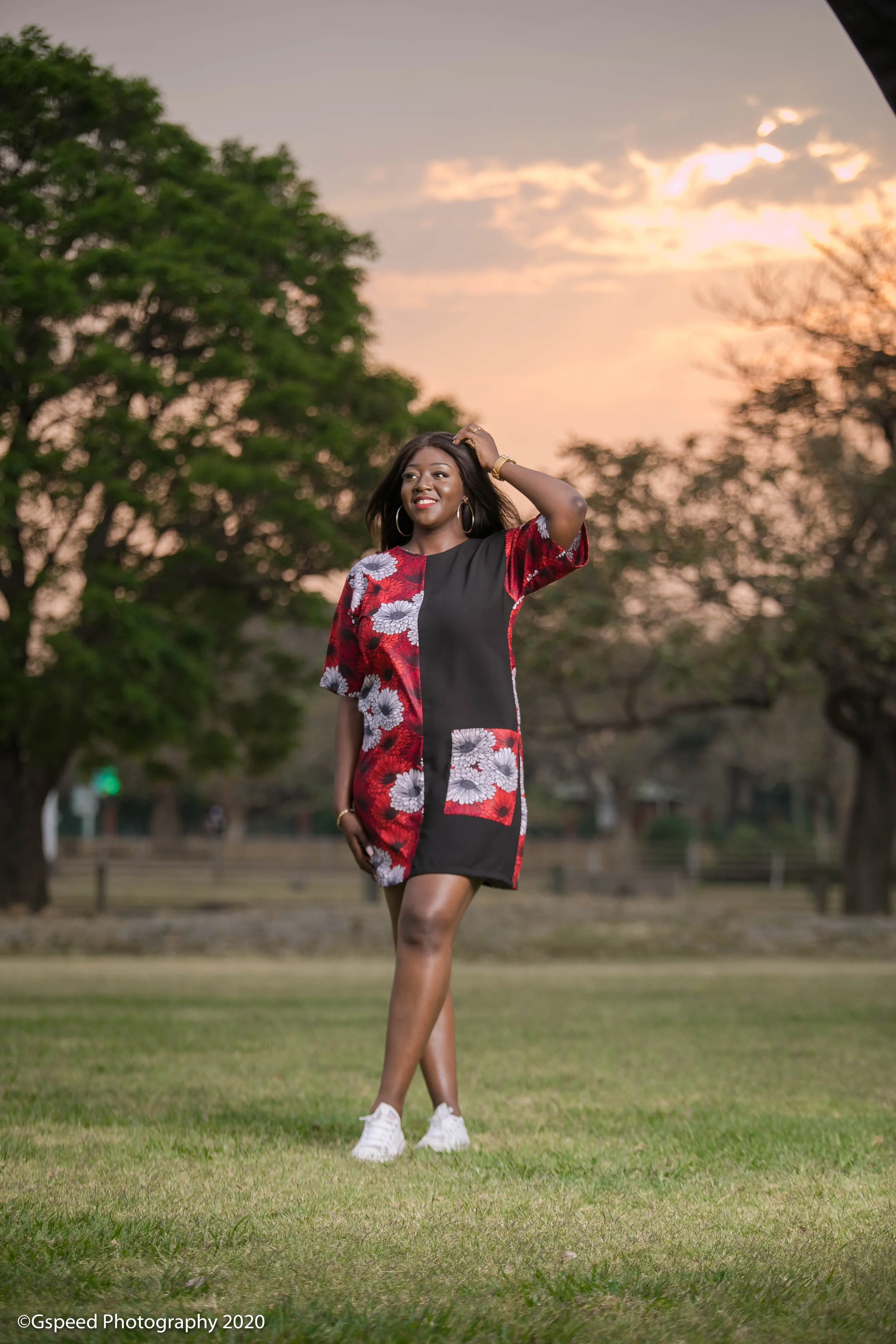 A woman in a black and red floral dress with white sneakers stands outdoors on a green lawn during sunset, with trees and a fence in the background.