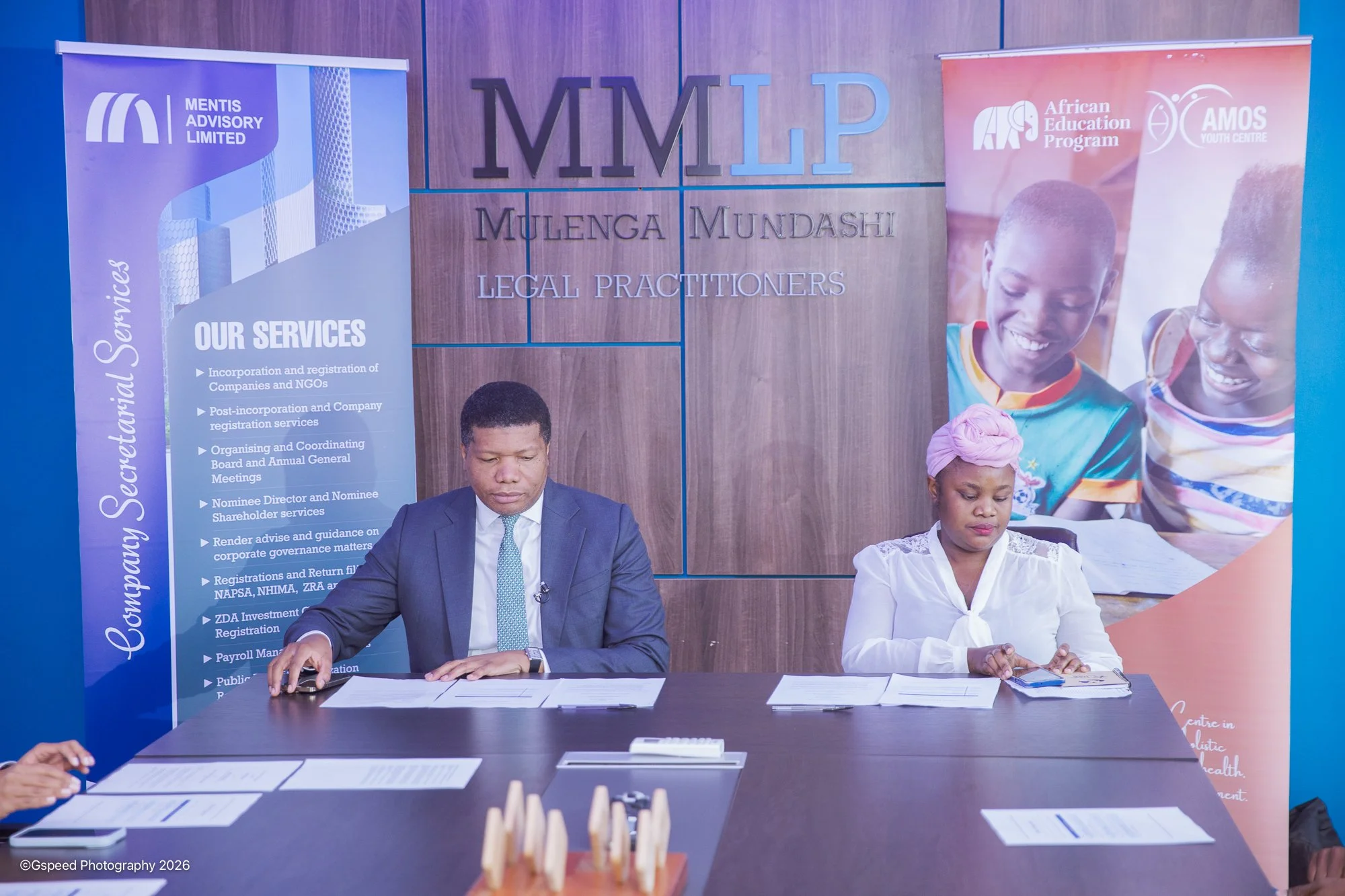 Two individuals sitting at a table in front of a wall with signs and banners for MMLP, Mentis Advisory Limited, and African Education Program. The man on the left is wearing a suit and the woman on the right is wearing a white blouse with a pink head
