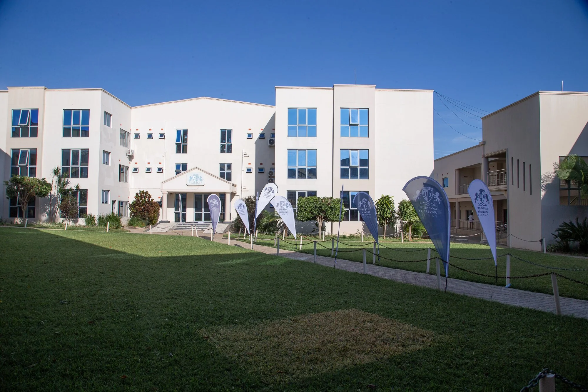 Modern white multi-story university building with large windows, green lawn, trees, banners, and clear blue sky.