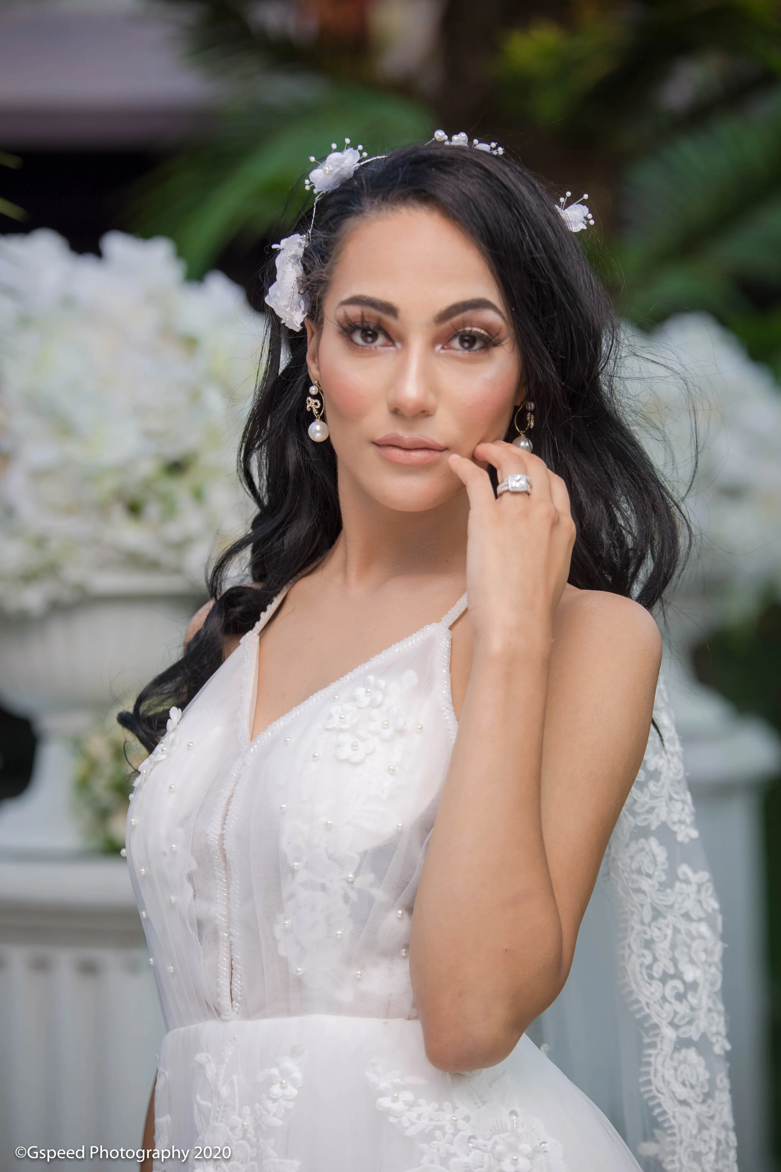 Woman in a white wedding dress with floral embroidery, wearing pearl earrings and a pearl and floral headpiece, posed outdoors with greenery and flowers in the background.