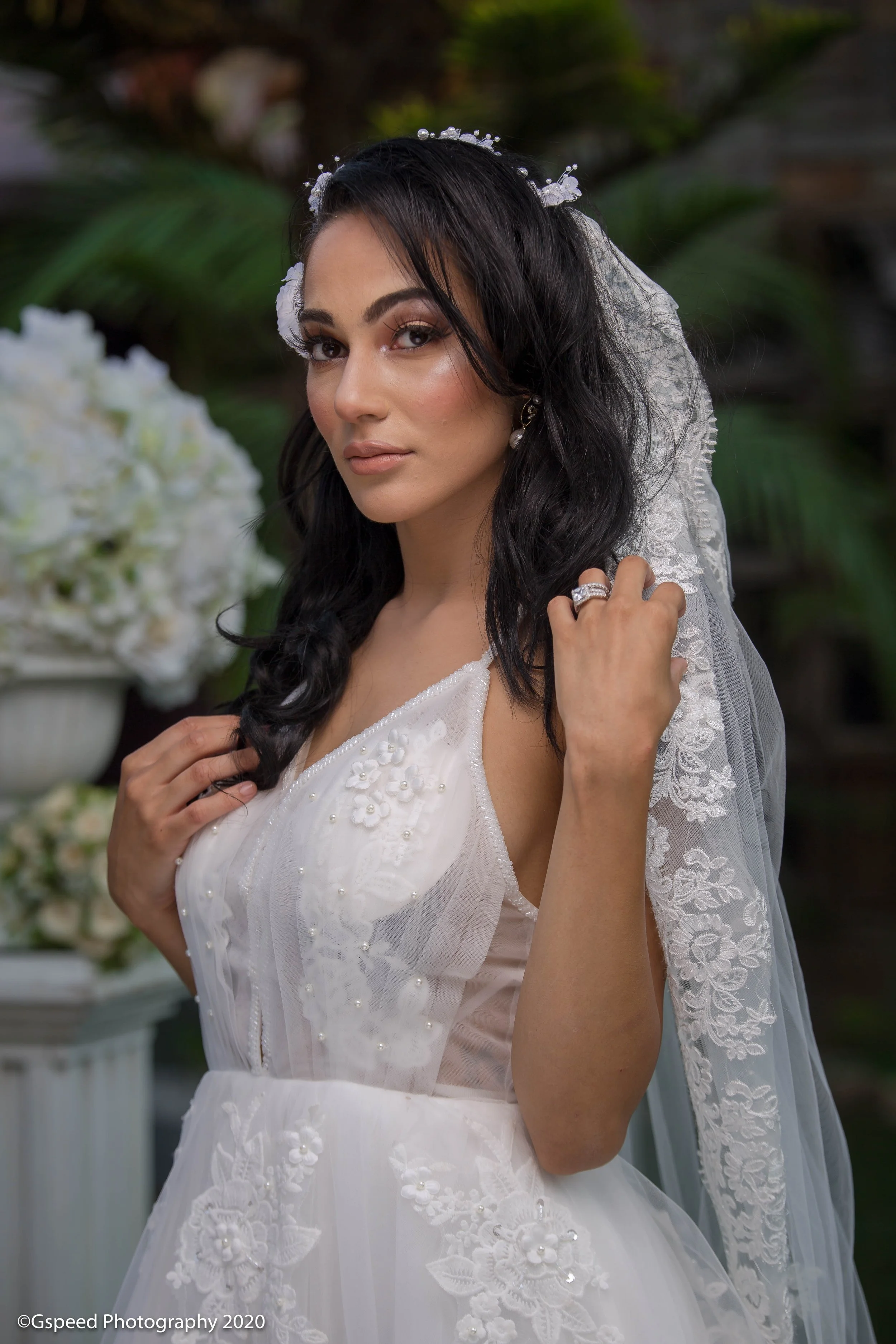 A bride in a white wedding dress with floral embroidery and pearls, black wavy hair, and a lace veil, outdoors with greenery and flowers in the background.
