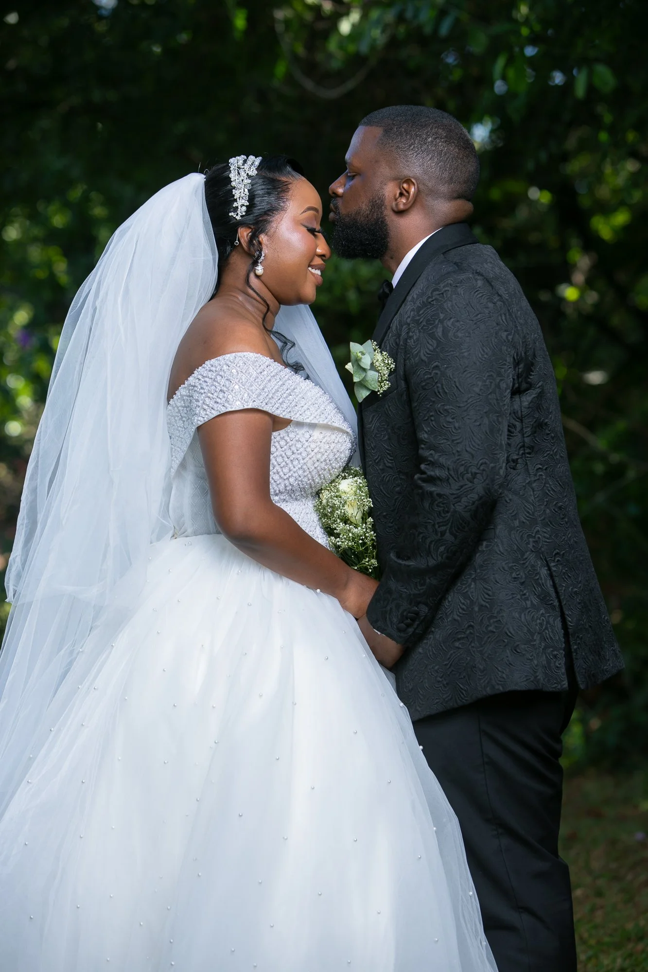 A bride and groom sharing a kiss outdoors, dressed in wedding attire, with green foliage in the background.
