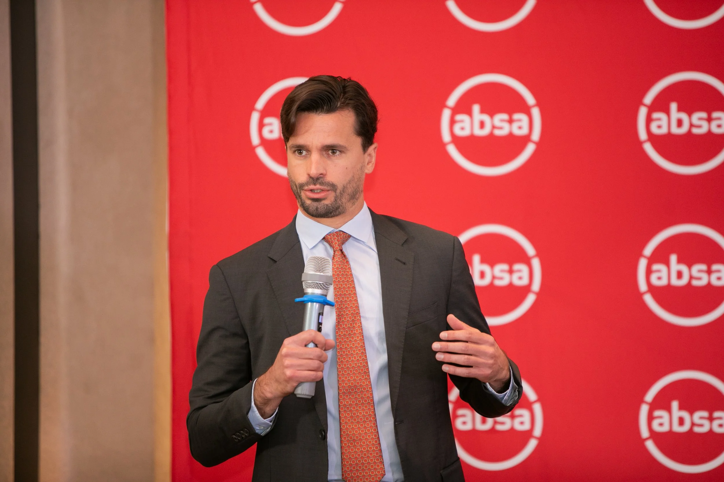 A man in a suit and tie holding a microphone, speaking at an event with a red backdrop featuring the 'absa' logo.