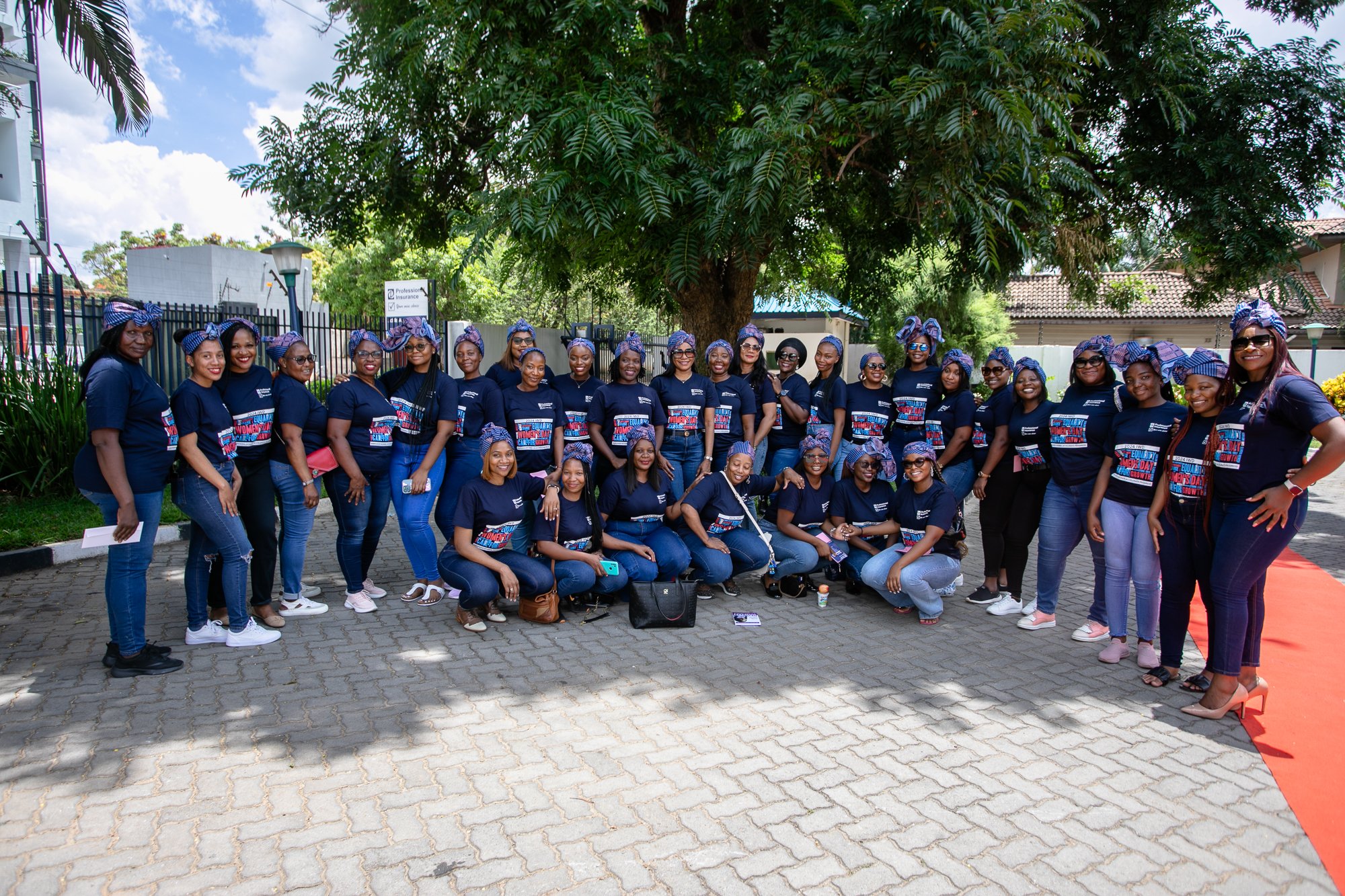 Group of women dressed in navy blue t-shirts and head wraps, standing outdoors in front of a large tree, posing for a photo during a daytime event that celebrates women's day.