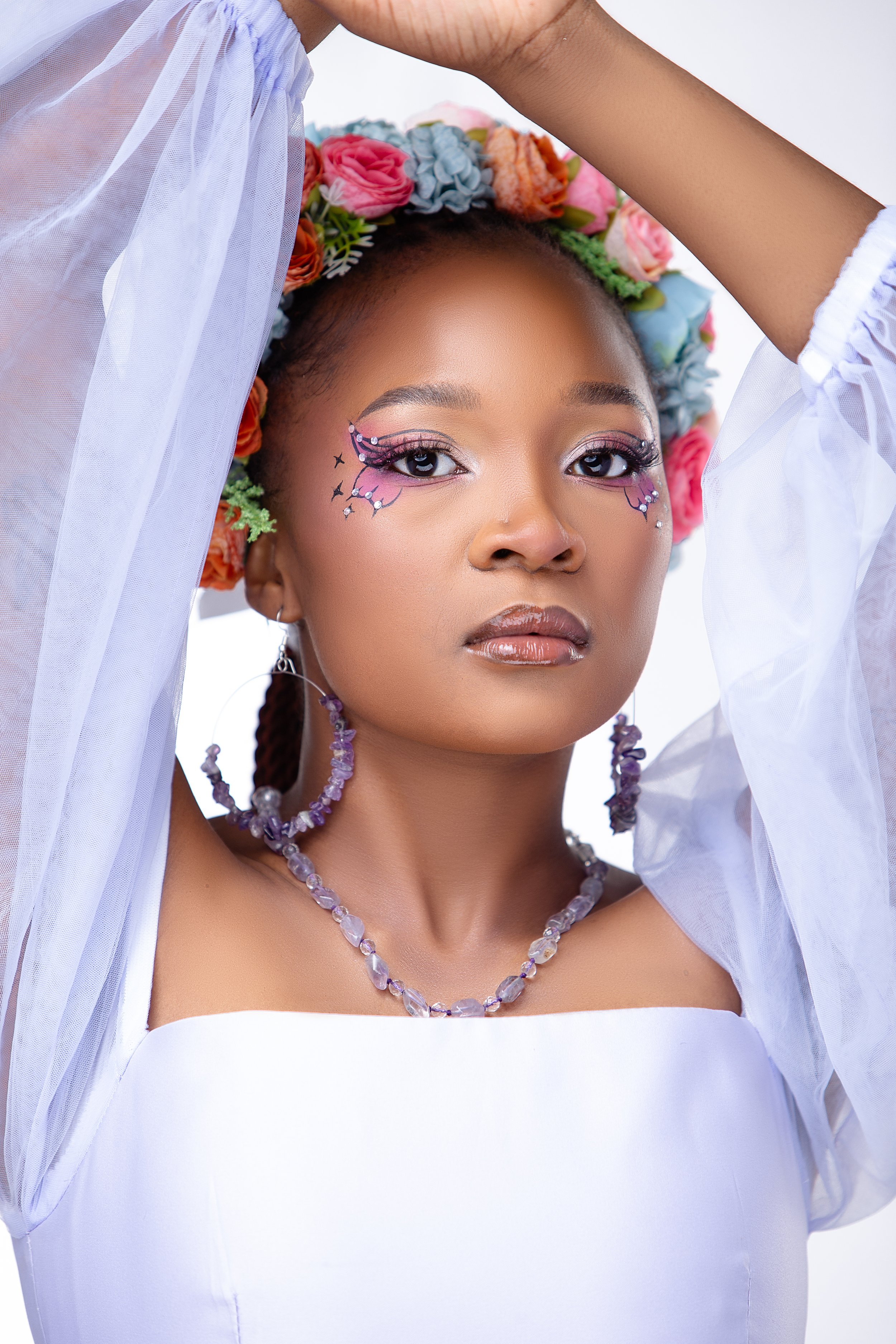 A woman with dark skin, styled in braids, is wearing a white off-shoulder top, and has elaborate eye makeup with butterfly and star designs, rhinestones, and pink eyeshadow. She is adorned with purple gemstone jewelry and a headband made of multicolo