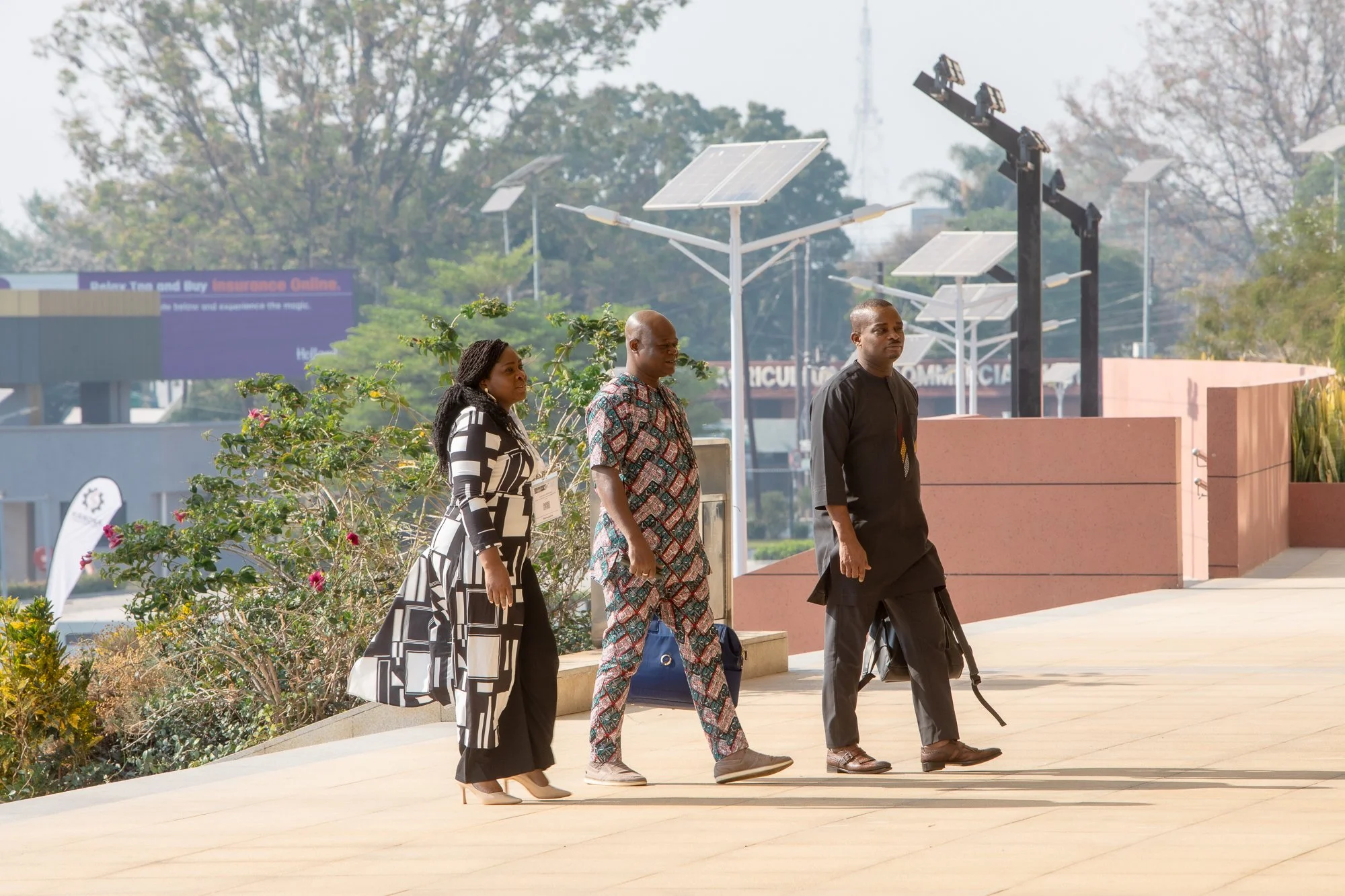 Three people walking outdoors on a sunny day near a pink wall and antenna equipment. One woman wears a black and white patterned dress, a man wears a colorful patterned shirt, and another man wears traditional black attire. There are solar panels on 