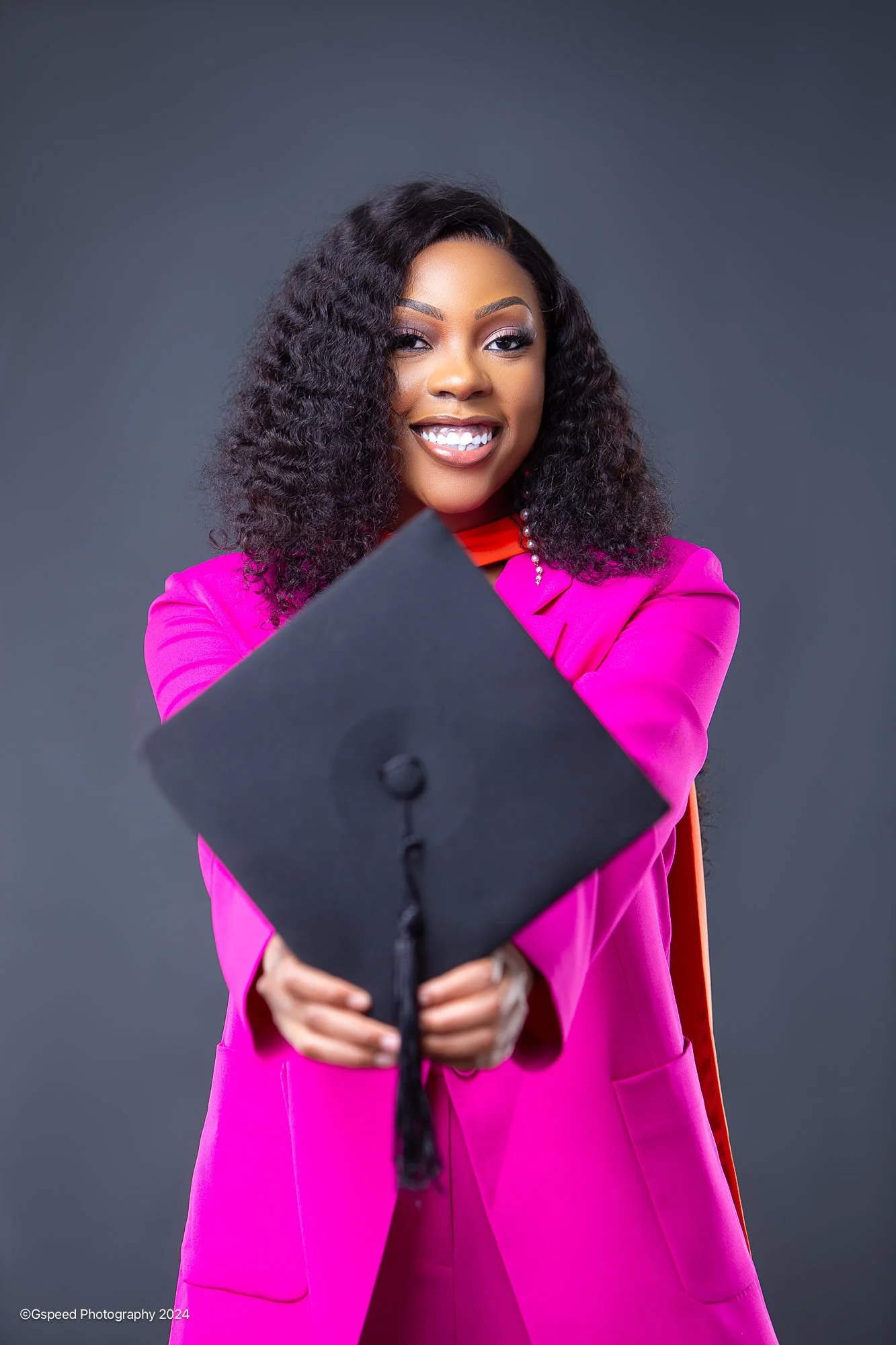 A smiling woman with curly black hair wearing a bright pink blazer and holding a graduation cap towards the camera.