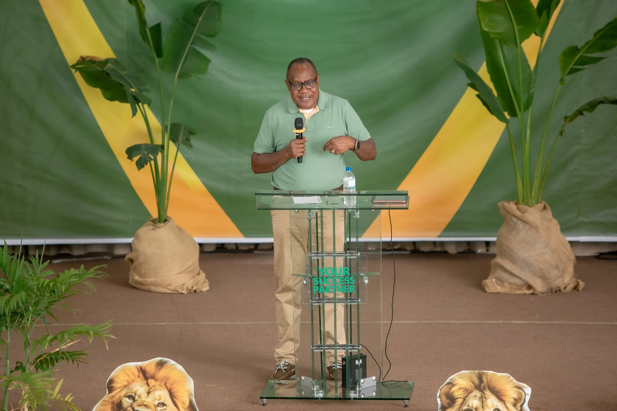 A man speaking at a transparent podium with a green read background, flanked by two large potted plants and lion cutouts on each side, with the words 'Your Success Partner' on the front of the podium.