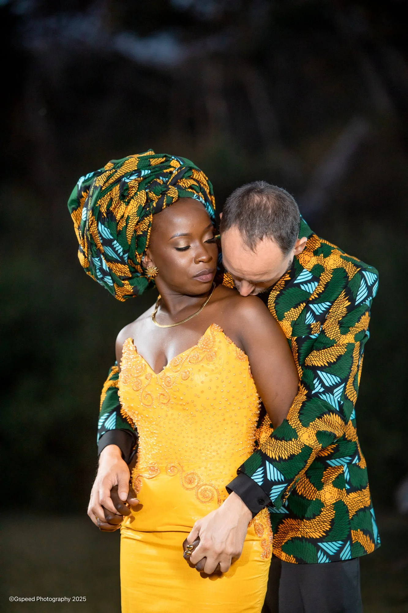 A woman in a bright yellow dress and a headwrap with an African print, and a man in a matching African print jacket, holding hands and embracing against a dark background, with the man kissing the woman's shoulder.
