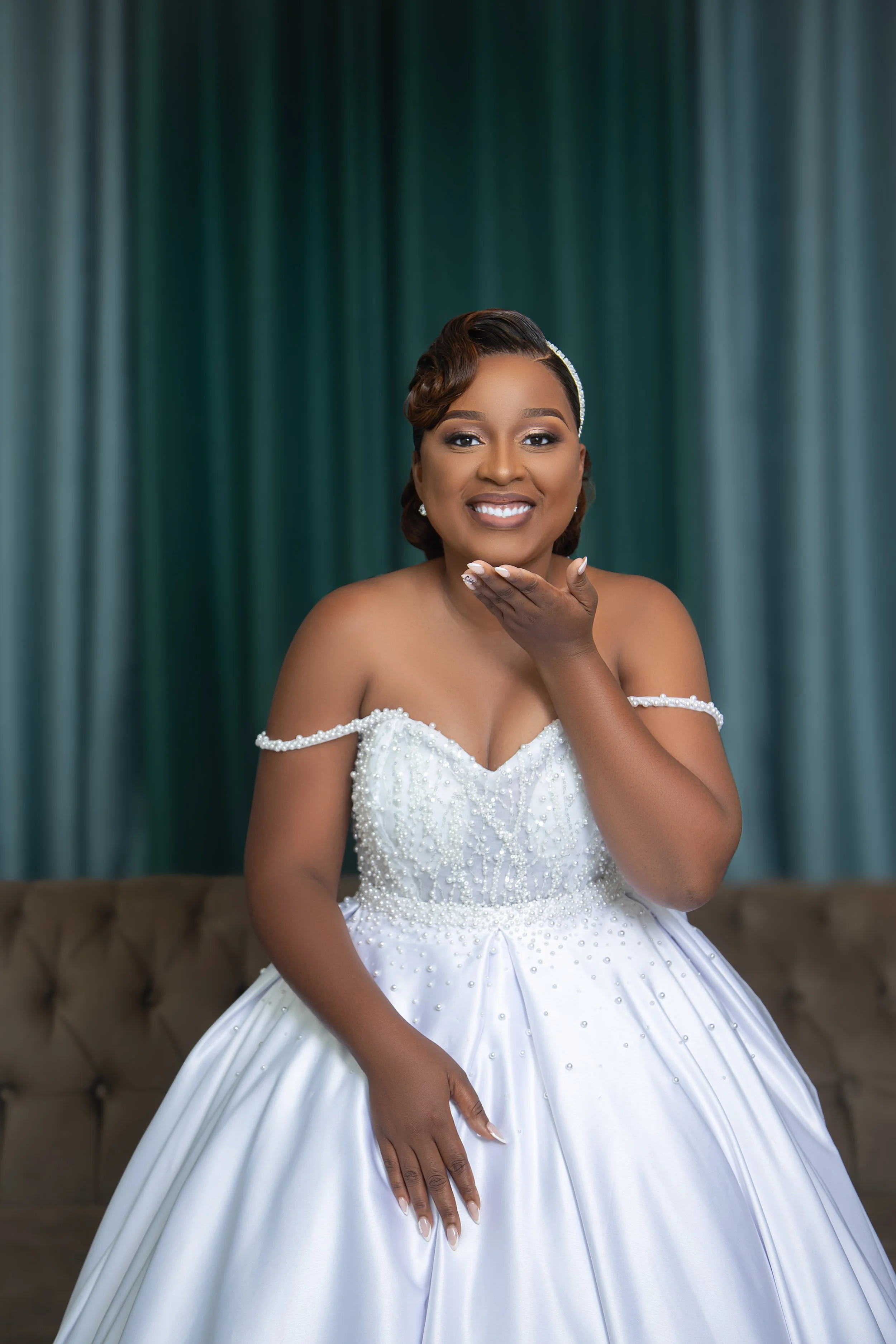 A smiling woman with brown hair styled in vintage waves, wearing a white wedding dress with pearl embellishments, posing in front of a green curtain backdrop.