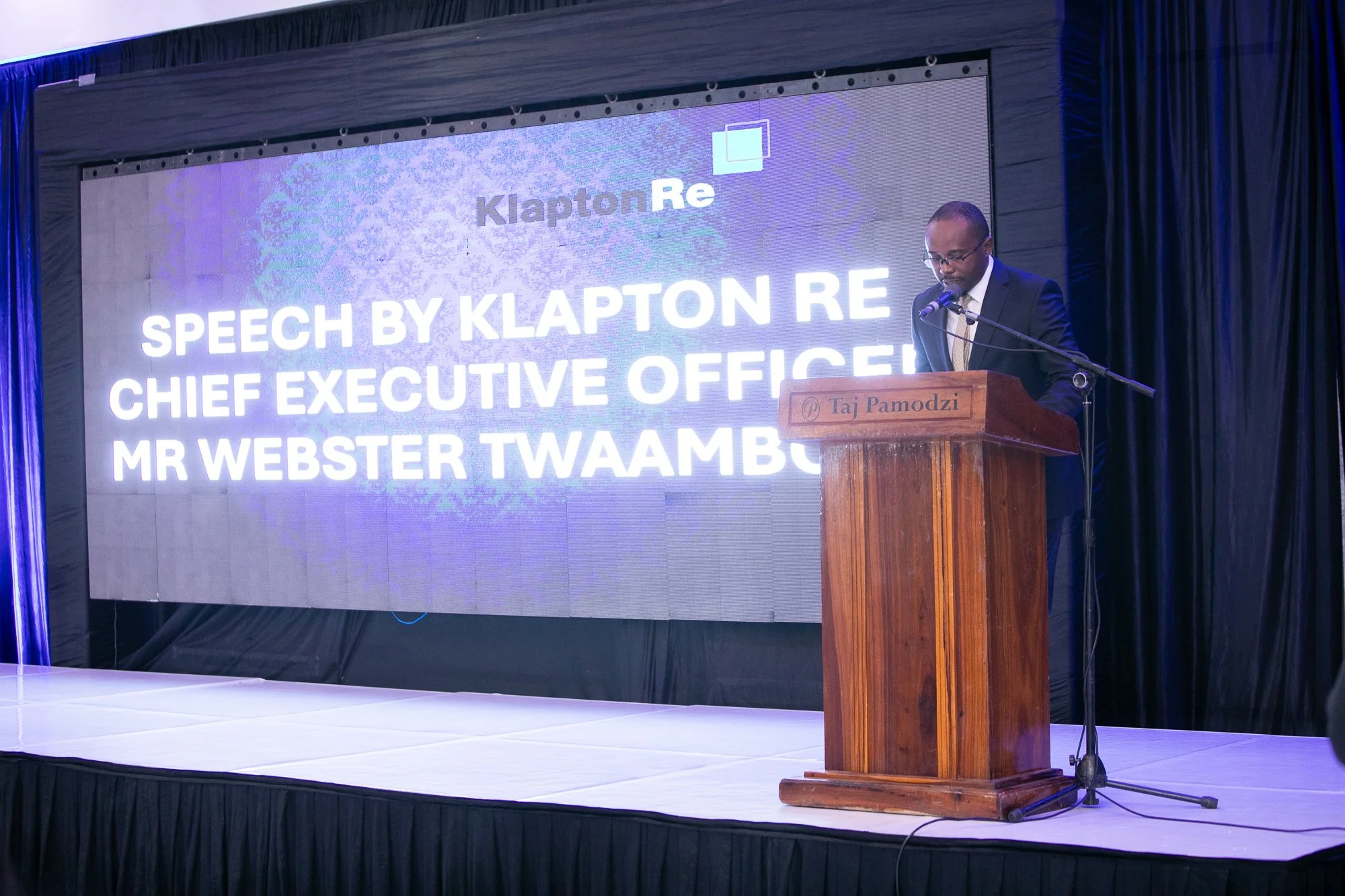 A man in a dark suit and glasses speaking at a wooden podium labeled 'Taj Pamodzi', with a microphone attached. Behind him, large screen displays white text about a speech by Klapton Re, the Chief Executive Officer, Mr. Webster Twaambo, at an event.
