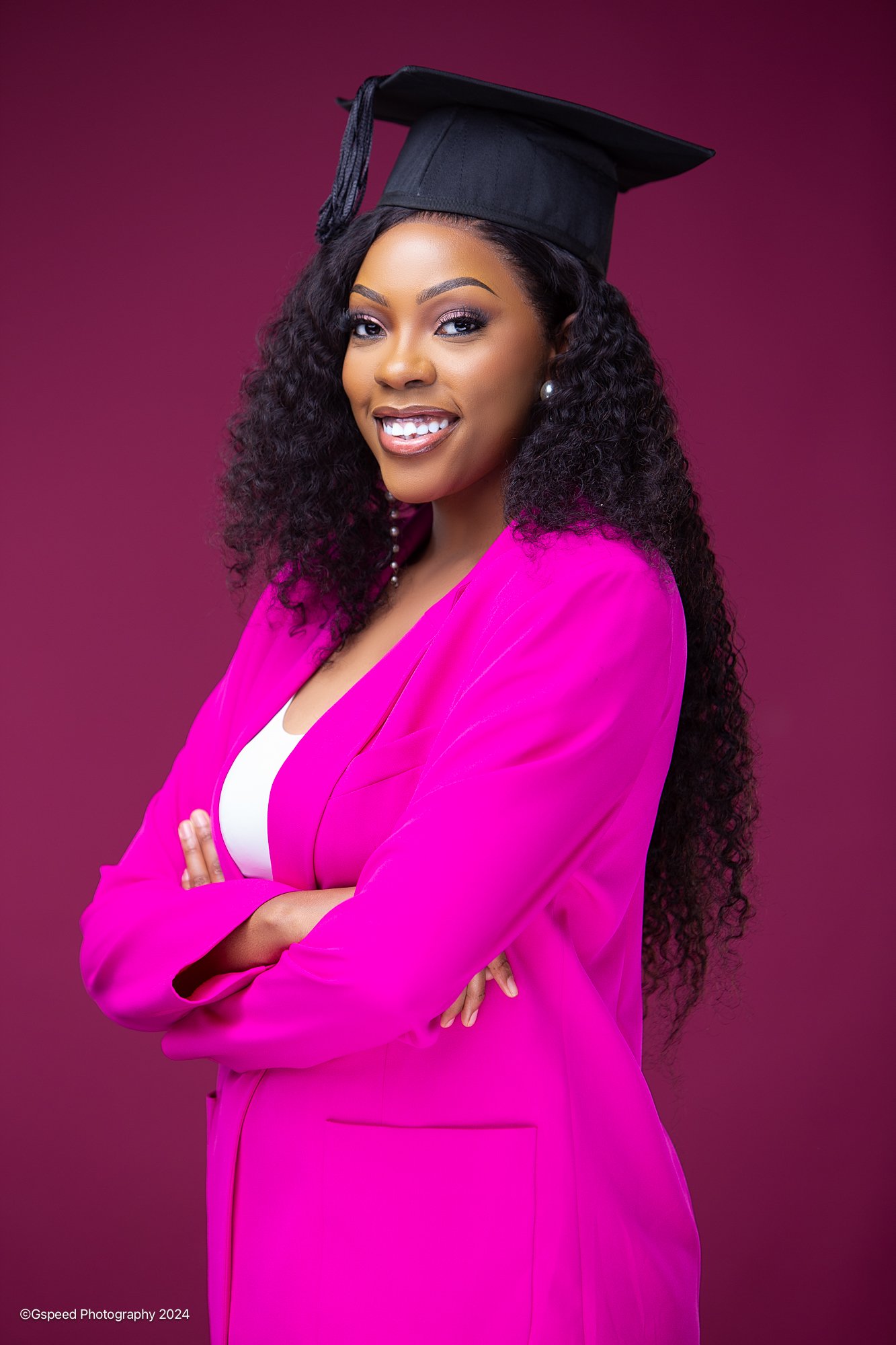 Portrait of a woman wearing a black graduation cap, bright pink blazer, crossing her arms, against a pink background.