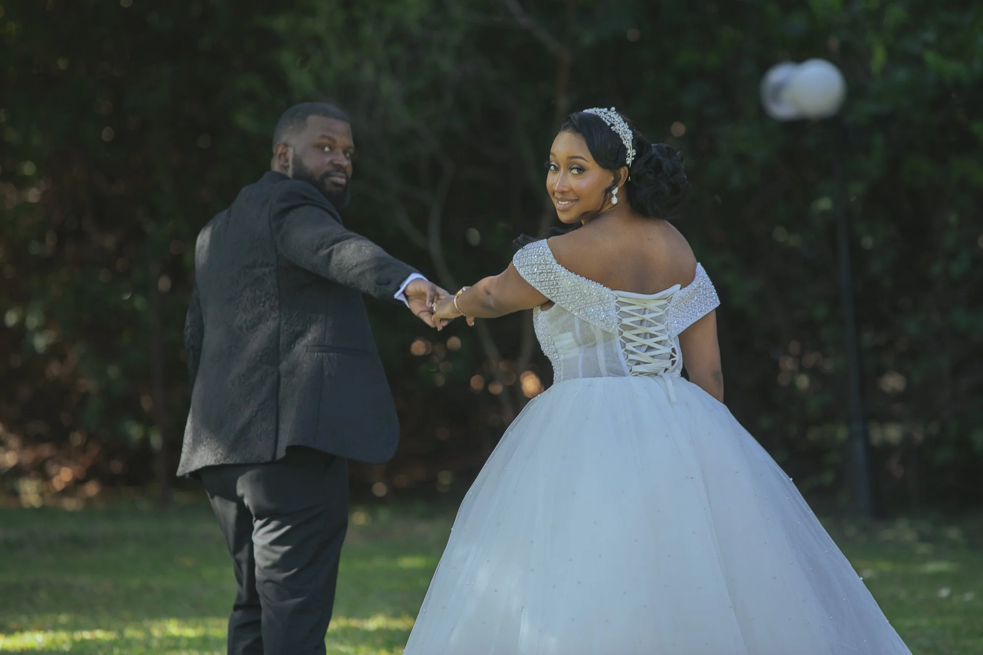 Couple dancing outside during wedding celebration, the woman wearing a white off-shoulder wedding dress with lace-up back and the man in a black suit, green background with trees.
