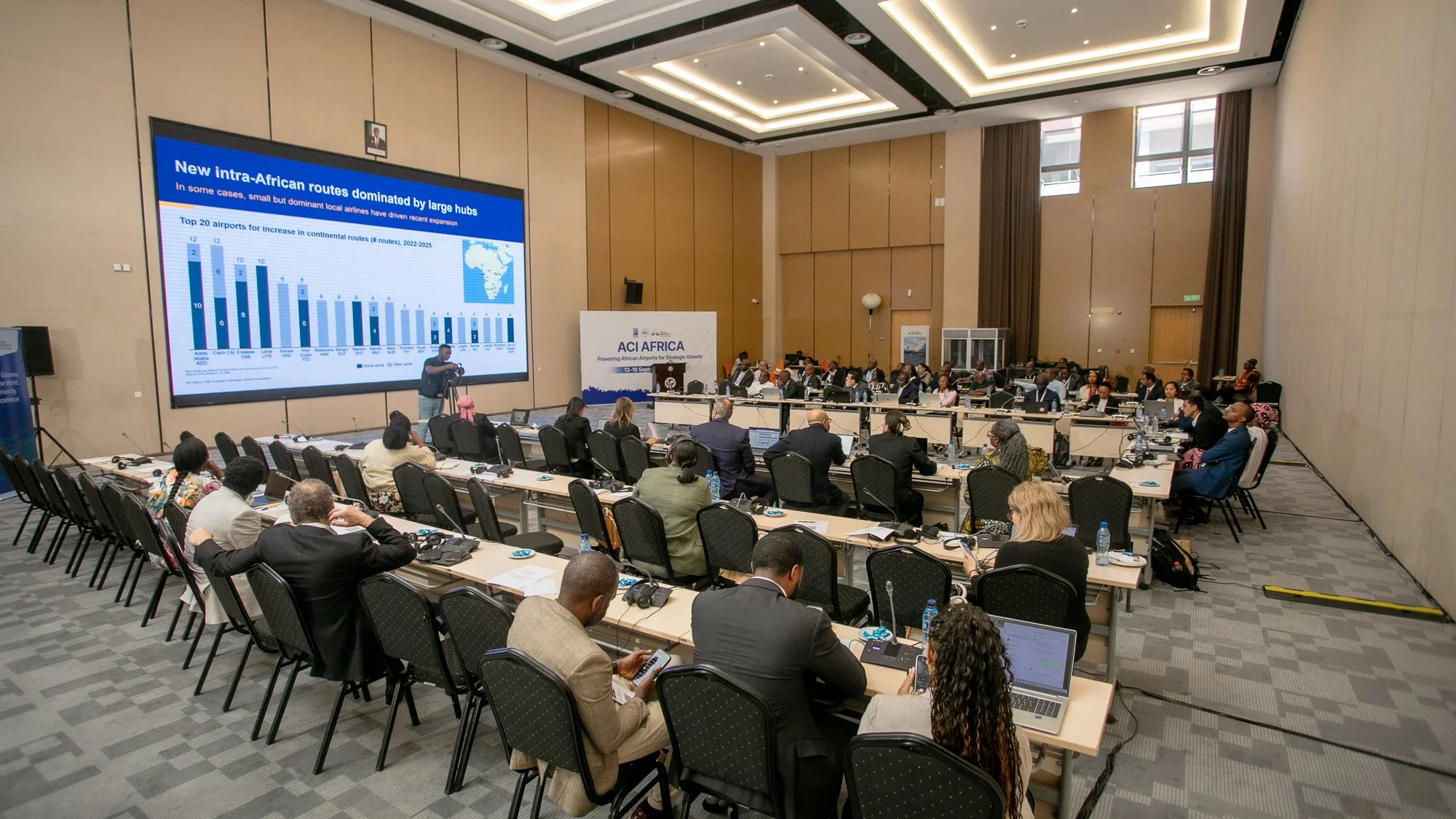 Conference room filled with seated attendees, a large digital screen displaying a presentation about African airline routes, and a speaker near the screen.