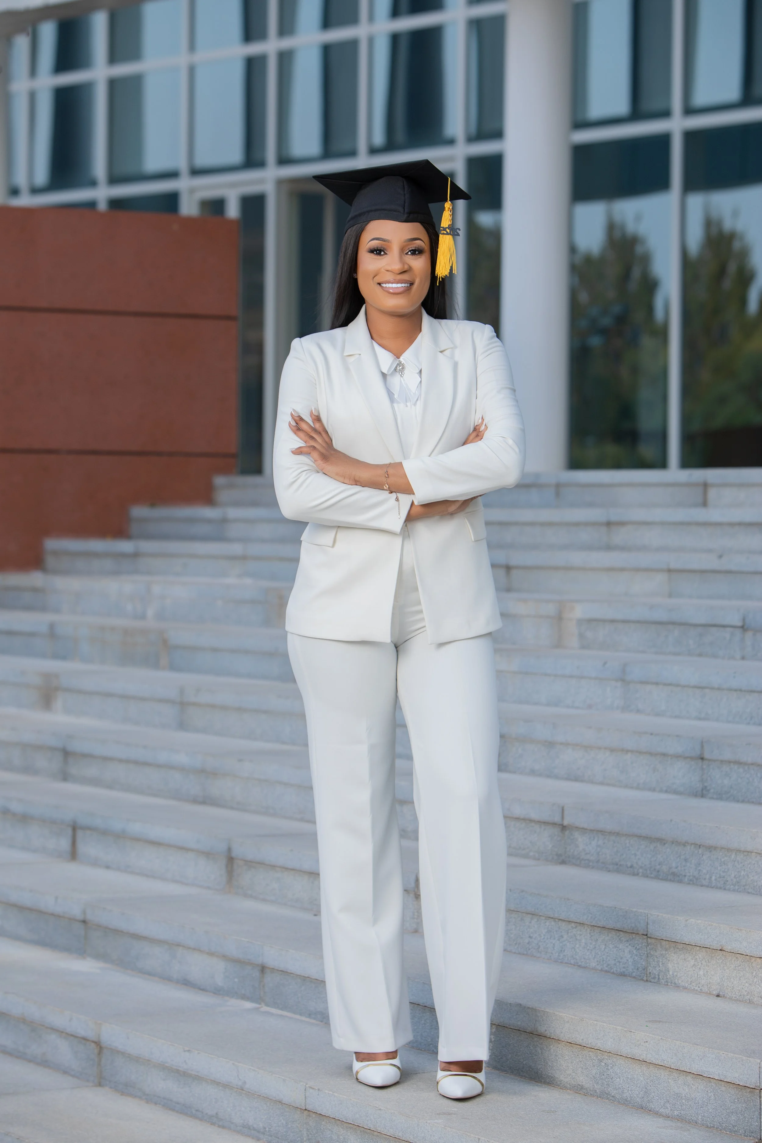 A young woman standing on concrete stairs outside a modern glass building, wearing a white suit, white shoes, and a black graduation cap with a yellow tassel, smiling with arms crossed.