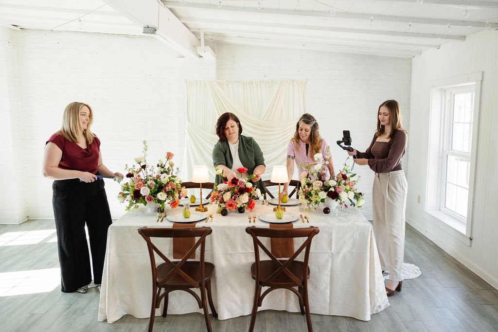 Four women, one holding a camera, preparing a decorated table with floral arrangements in a bright, white-room with natural light.