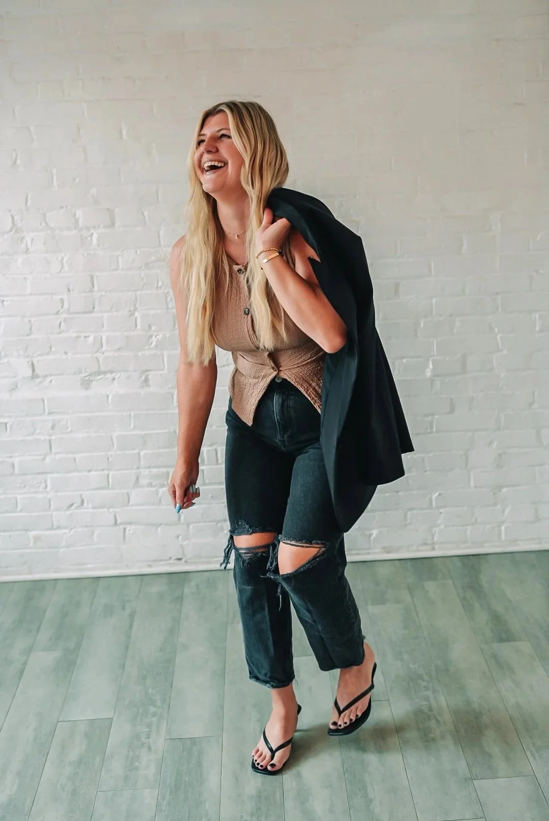 A woman with long blonde hair laughing while carrying a black blazer over her shoulder, wearing a sleeveless beige top, ripped black jeans, and black high-heeled sandals, standing against a white brick wall.