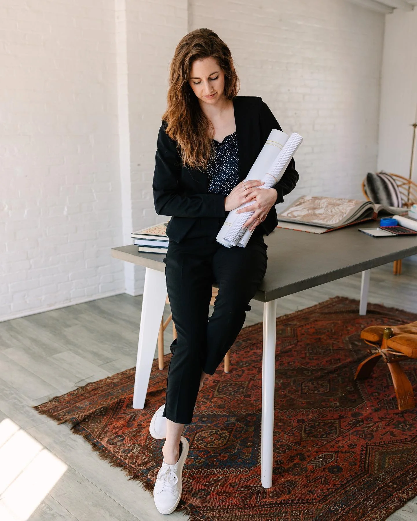 woman sitting on edge of desk, looking down, as she holds onto rolls of blueprints