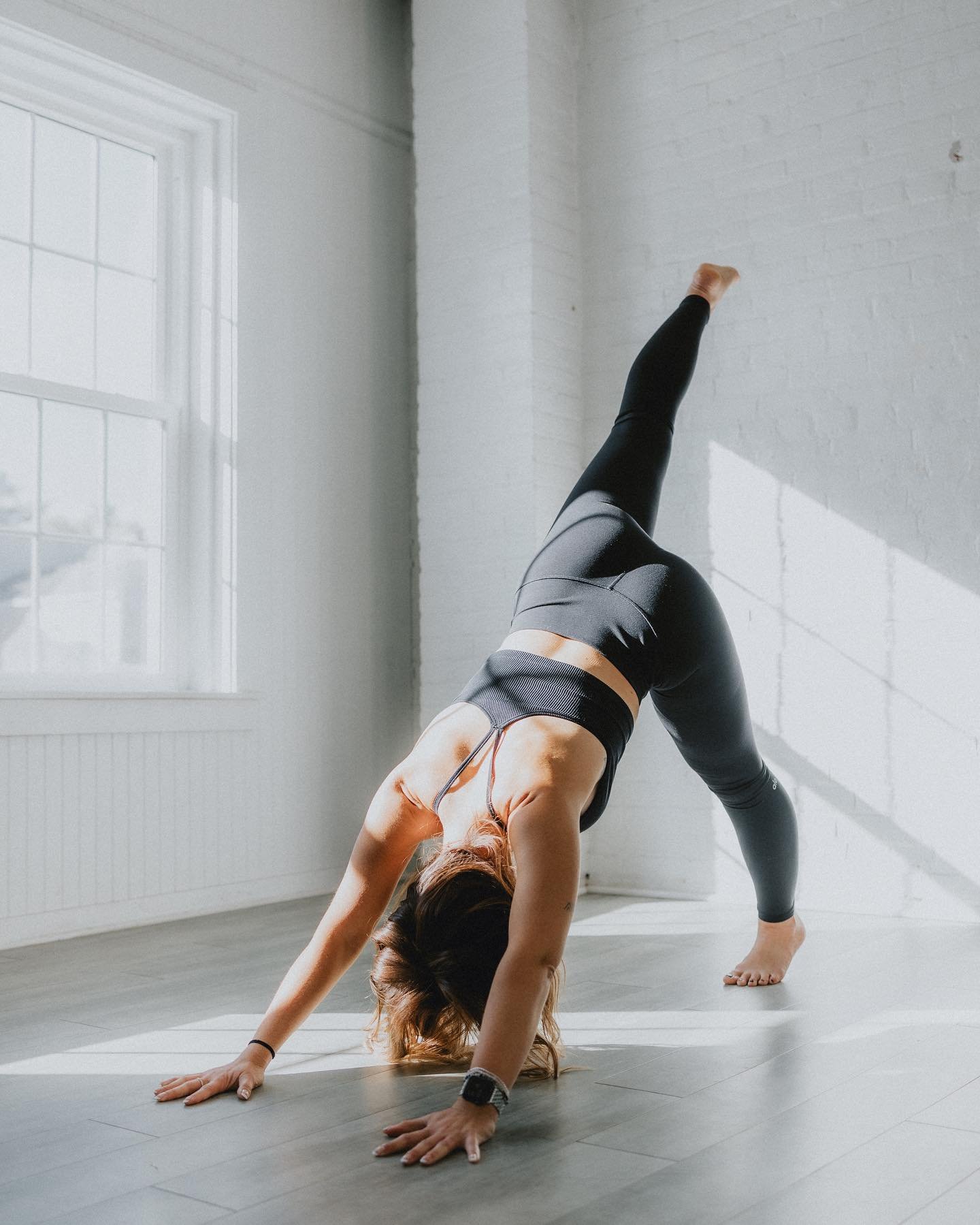 woman doing downward dog yoga pose with her right foot raised high into the air by white brick wall and window