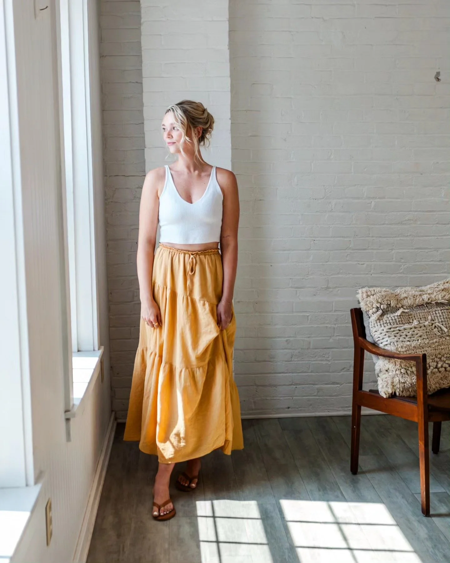 woman standing beside and looking out window in white tank top and long yellow skirt
