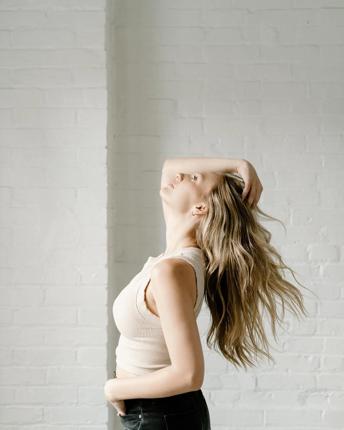 woman facing bright light of window as she leans way back and combs through her hair by white brick wall