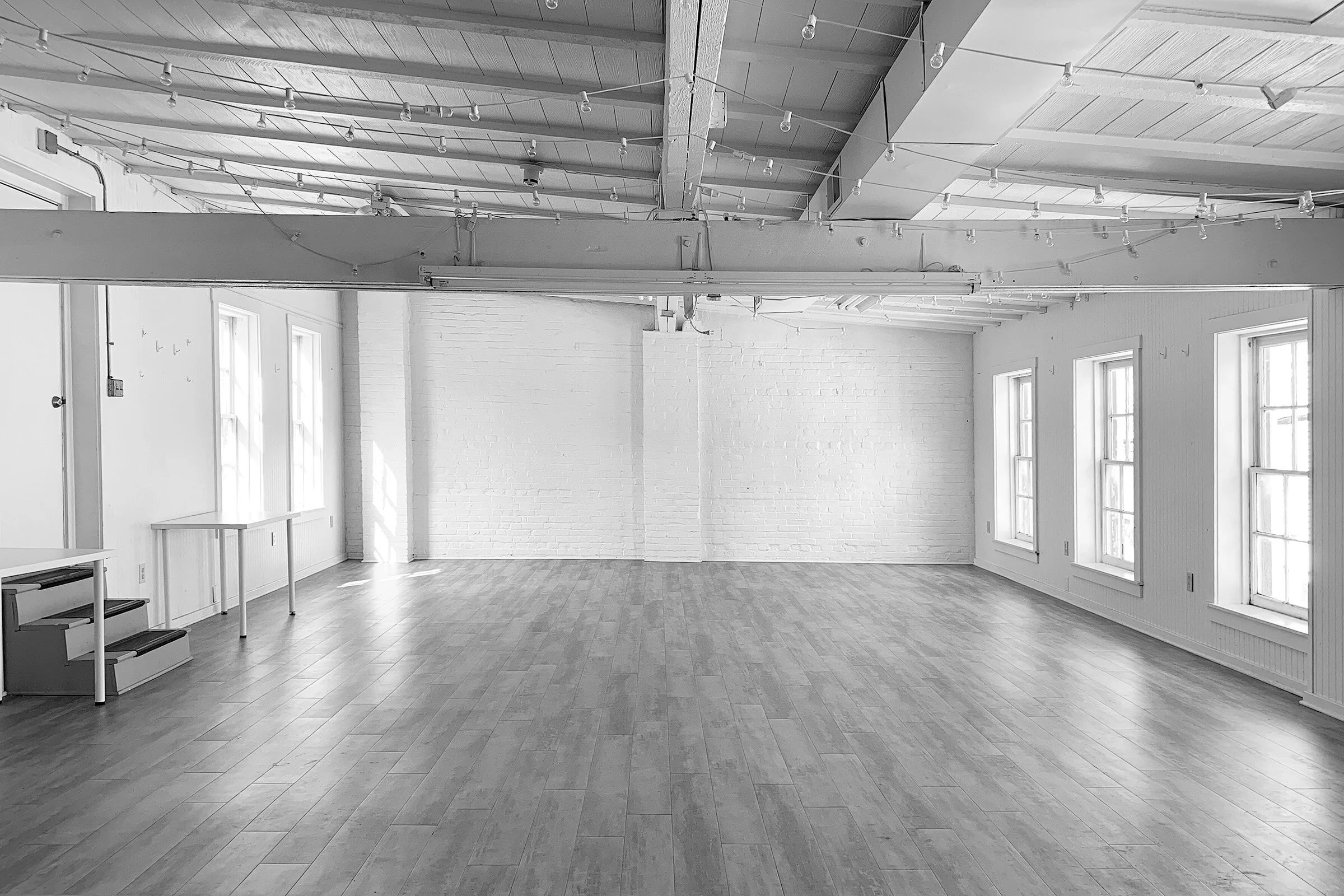 An empty room with large windows, light-colored wooden floors, and white brick walls. There are several large windows along one wall and string lights hanging from the ceiling. A white table and a set of small stairs are also visible.