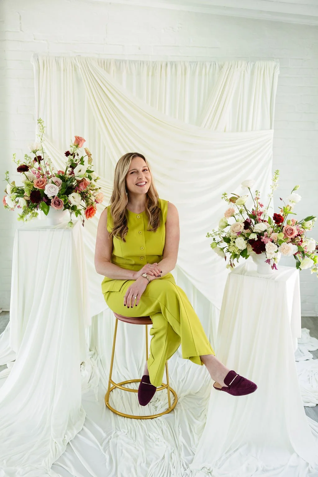 woman sitting on stool by white curtains and floral arrangements for brand shoot