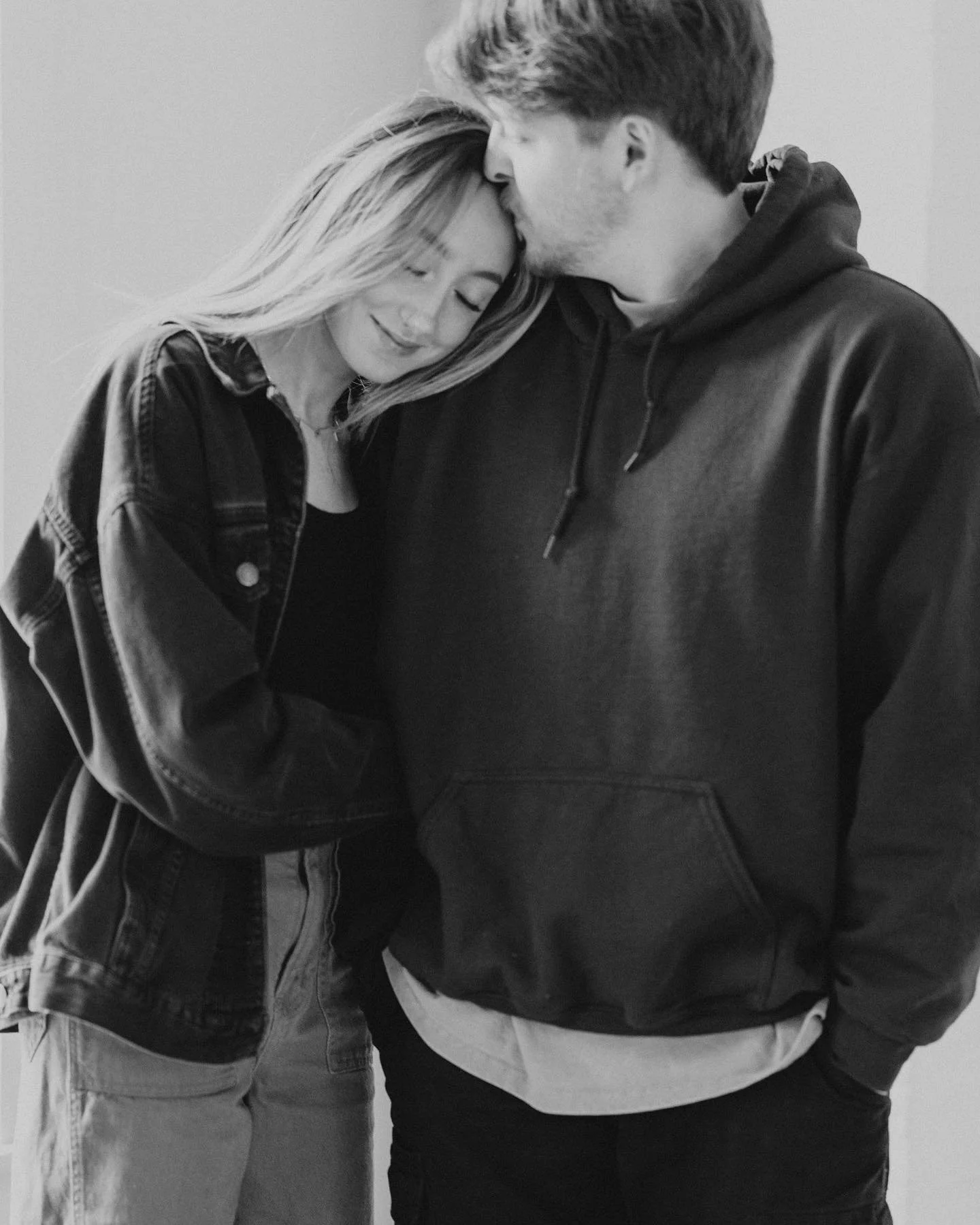 close up black and white photo of woman resting her left cheek on man's right shoulder as he kisses the top of her head