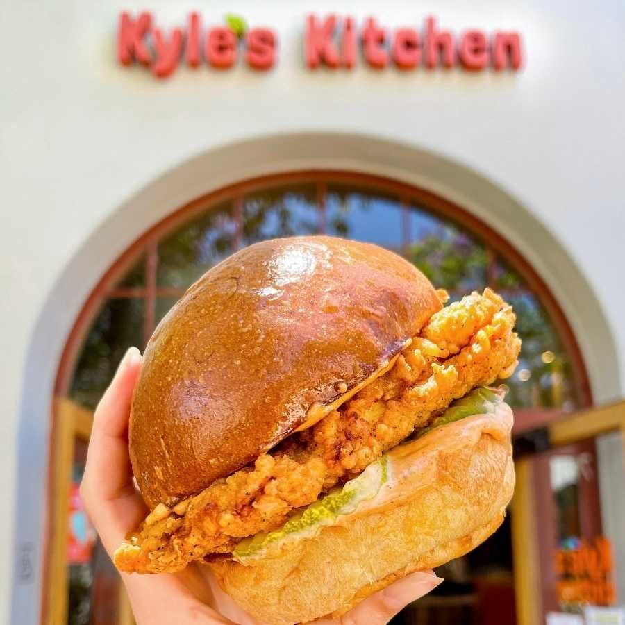 www.santabarbarawedding.com | Kyle’s Kitchen | Fried Chicken Sandwich Held in Front of the Kyle’s Kitchen Storefront