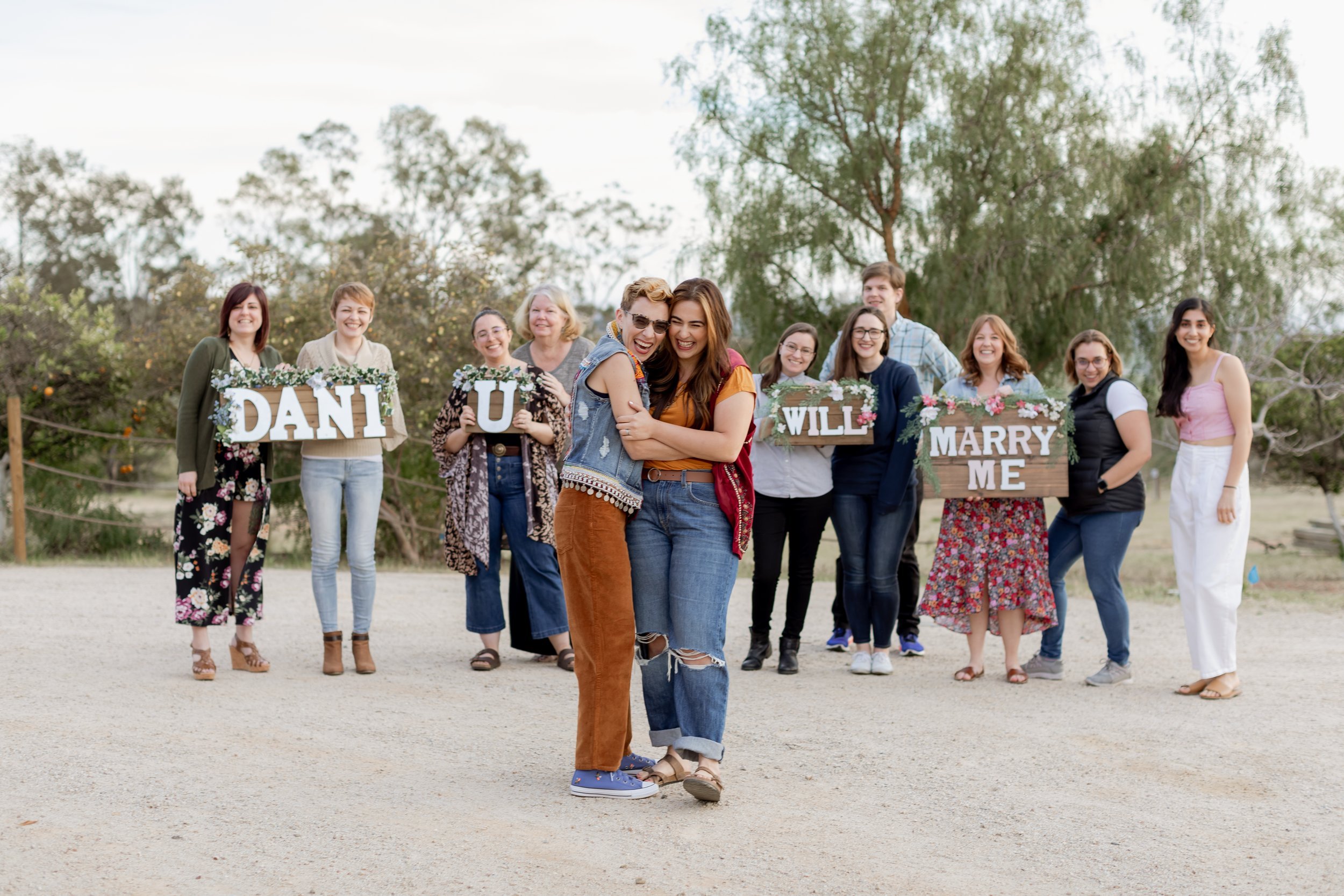 www.santabarbarawedding.com | MacKenzie Rana Photography | Fish House San Luis Obispo | Couple Embracing in Front of Friends and Family with Signs at Engagement Shoot