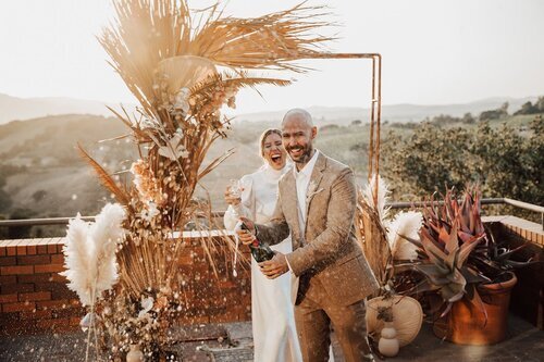 www.santabarbarawedding.com | The Dalarus Photography | Boho Chic Dreams | Couple Popping Champagne in Front of Wedding Arch with Unique Florals