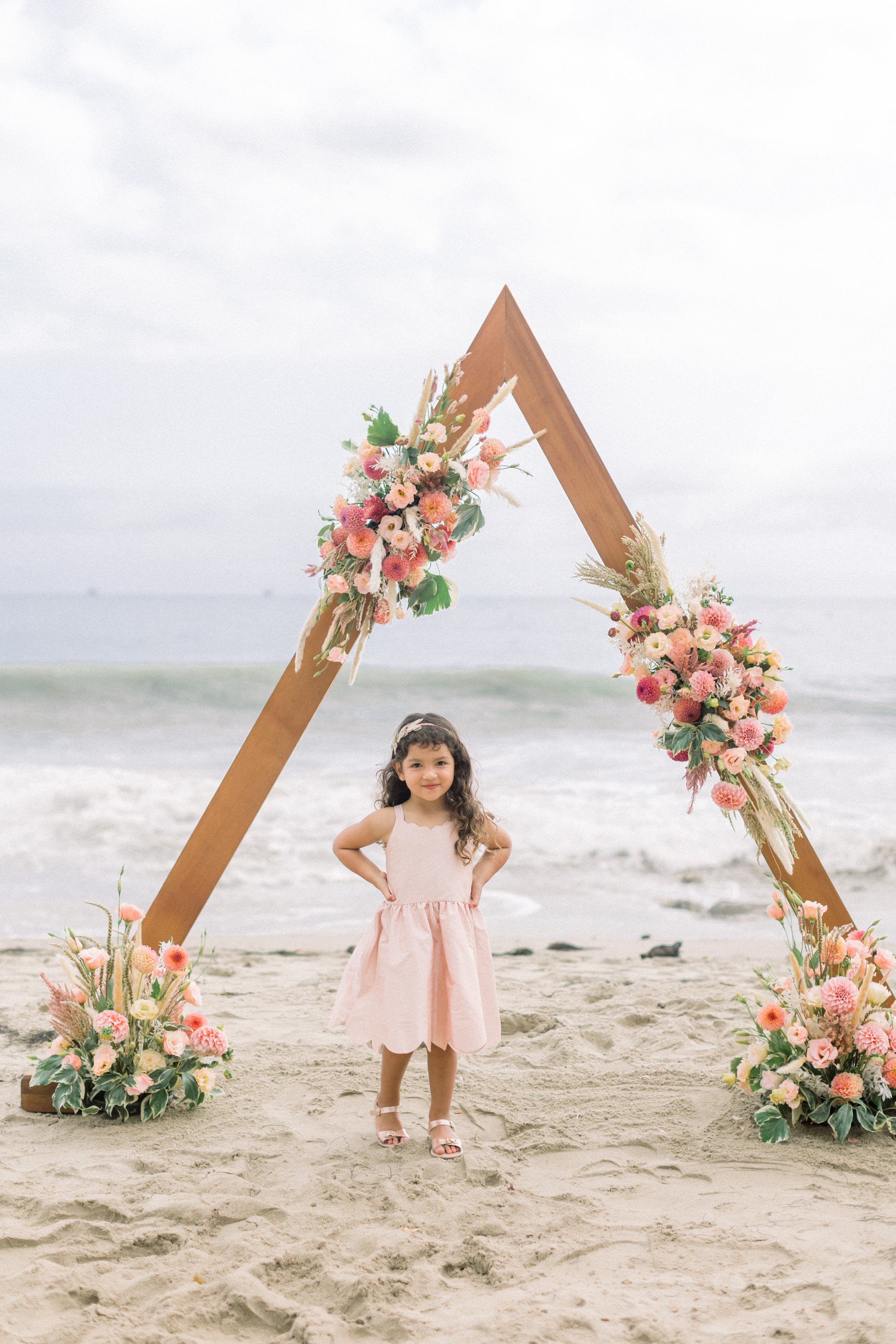 www.santabarbarawedding.com | James &amp; Jess Photography | Slate Catering | All Heart Rentals | Ella &amp; Louie | Flower Girl Standing in Front of Triangular Shaped Wedding Arch 