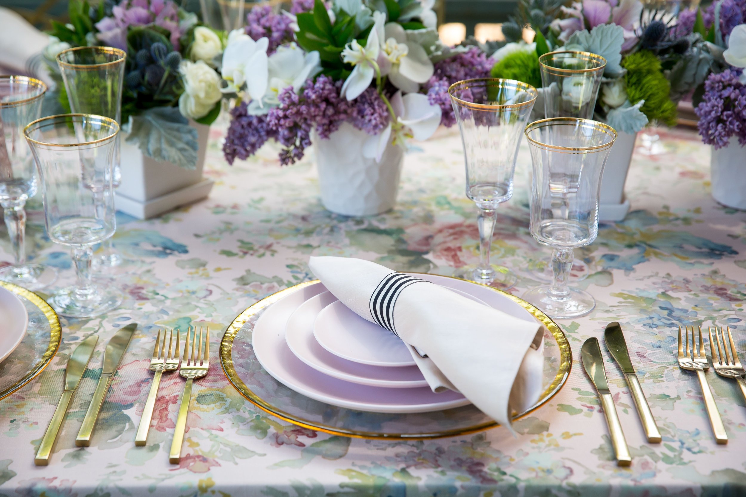 www.santabarbarawedding.com | Bright Event Rentals | Vanity Portrait Studio | Place Setting with Light Purple Plates, Gold Rimmed Glasses, and Purple Florals All on a Floral Tablecloth