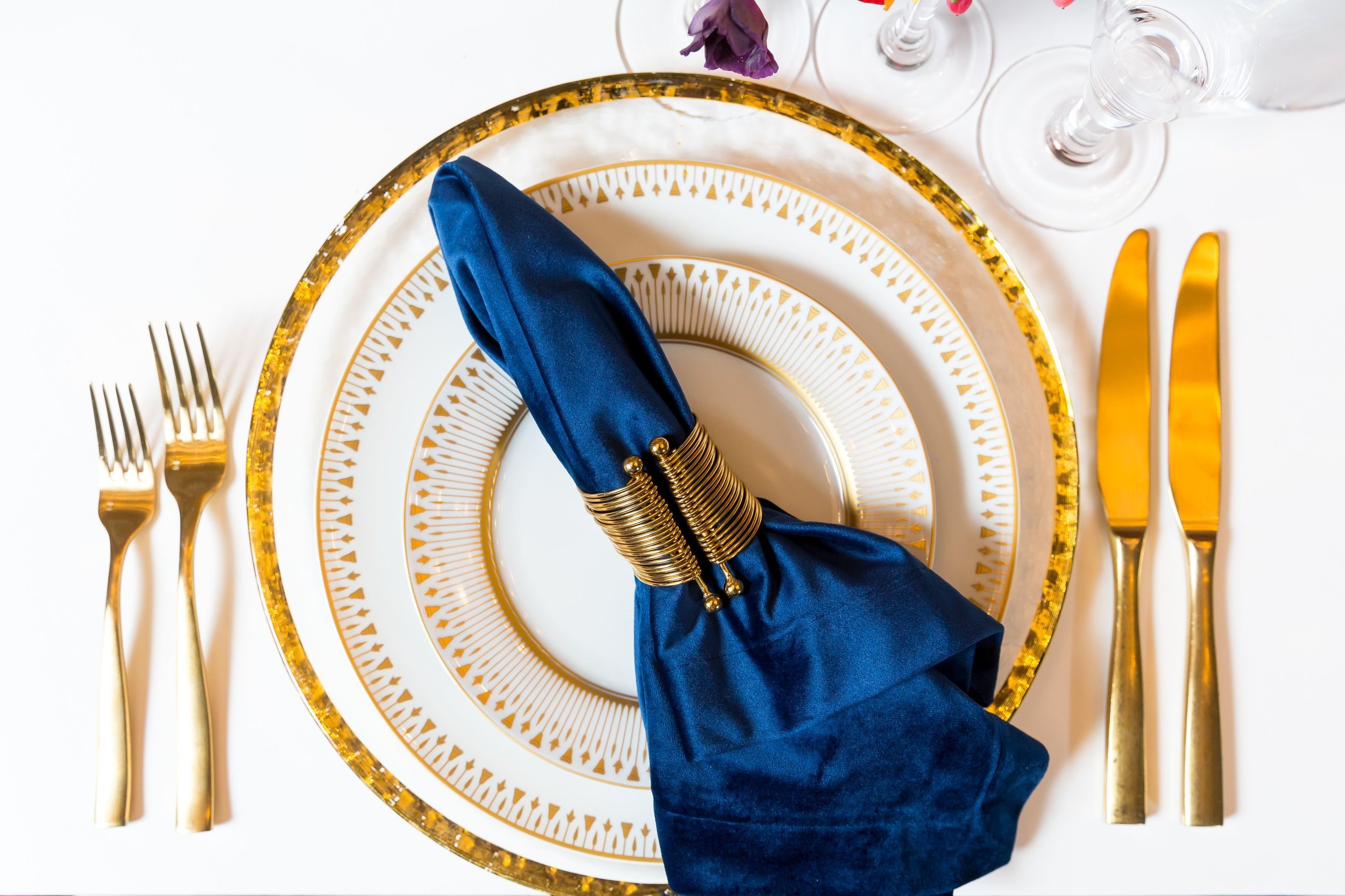 www.santabarbarawedding.com | Bright Event Rentals | Vanity Portrait Studio | Place Setting with Gold Rimmed Plates and Charger, Gold Silverware, and a Blue Napkin
