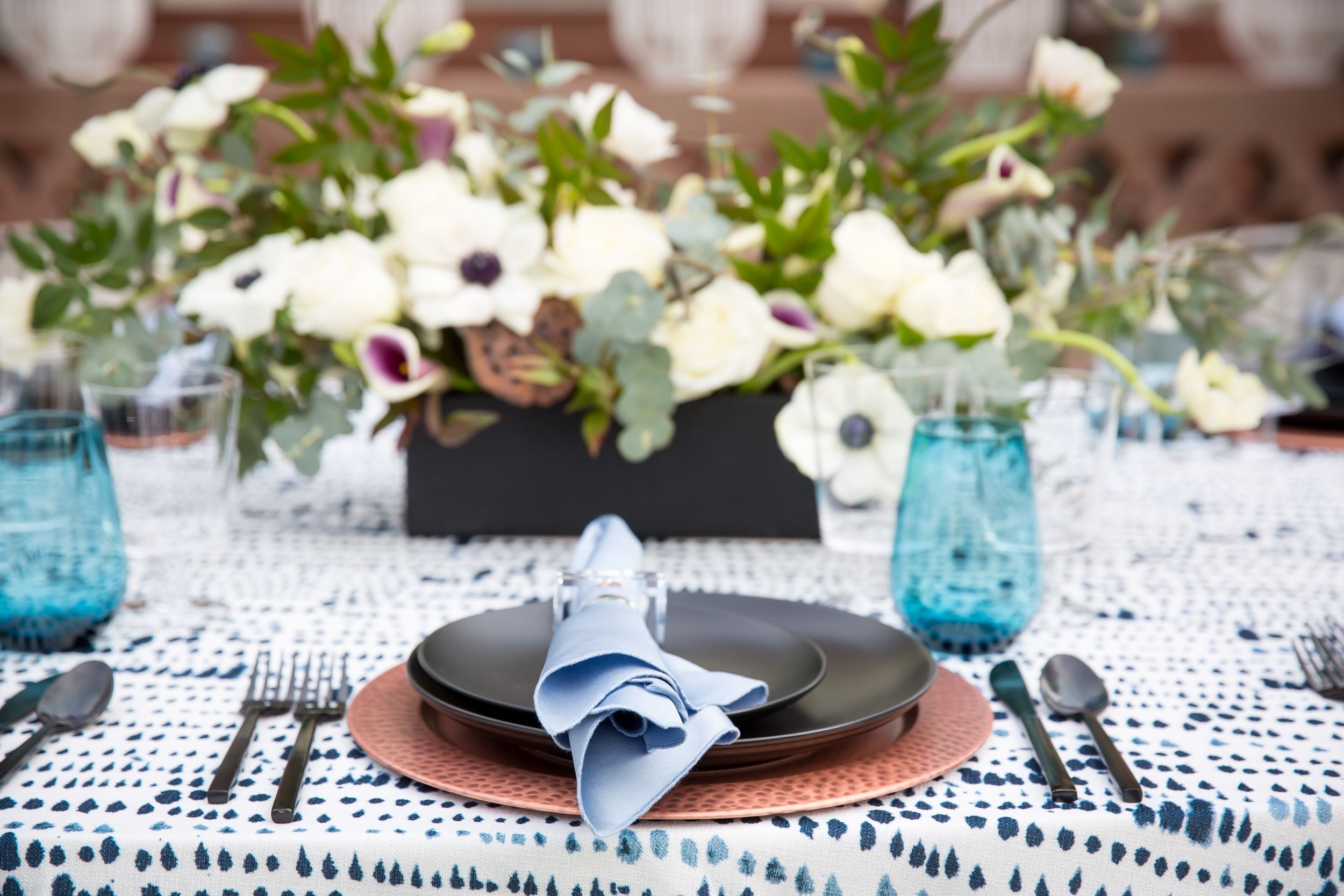 www.santabarbarawedding.com | Bright Event Rentals | Vanity Portrait Studio | Place Setting with Black Plates on a Brown Charger, Blue Glasses and Napkins All on a Blue and White Tablecloth