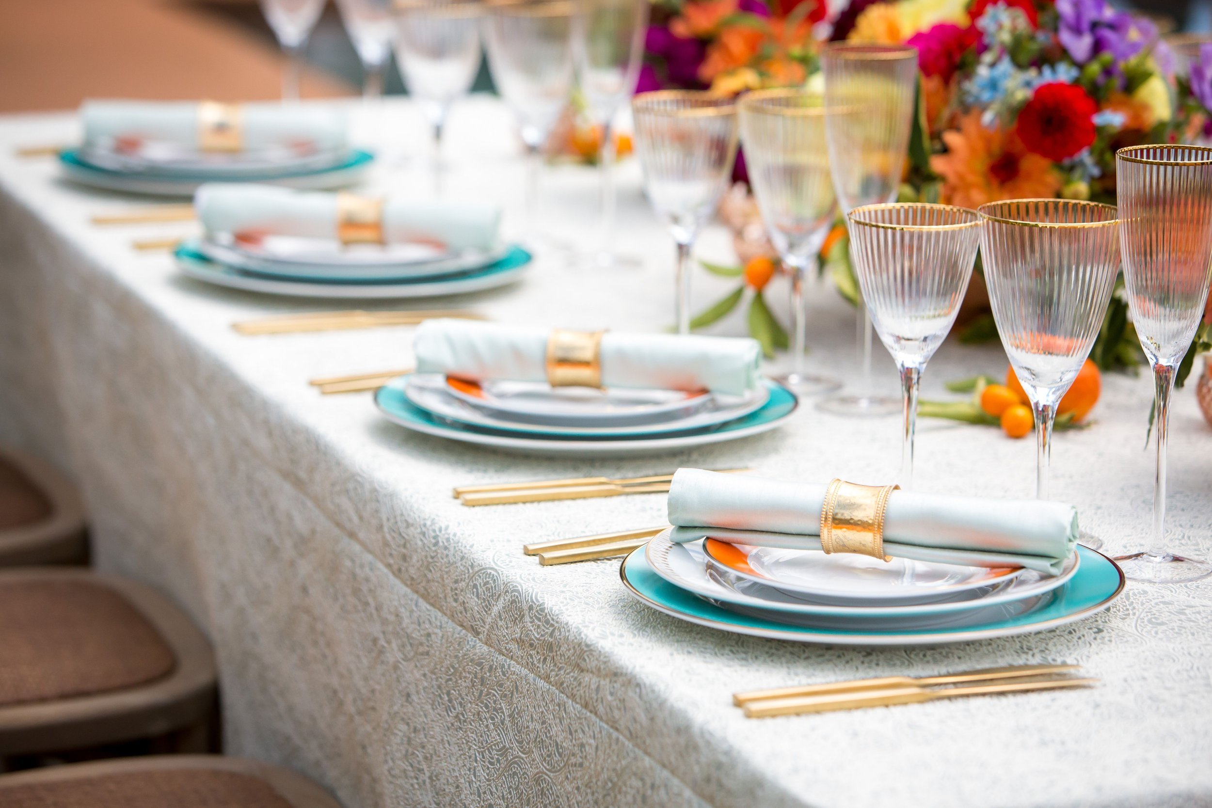 www.santabarbarawedding.com | Bright Event Rentals | Vanity Portrait Studio | Place Settings with Blue, White, and Orange Plates, Gold Silverware, and Gold-Rimmed Stemware