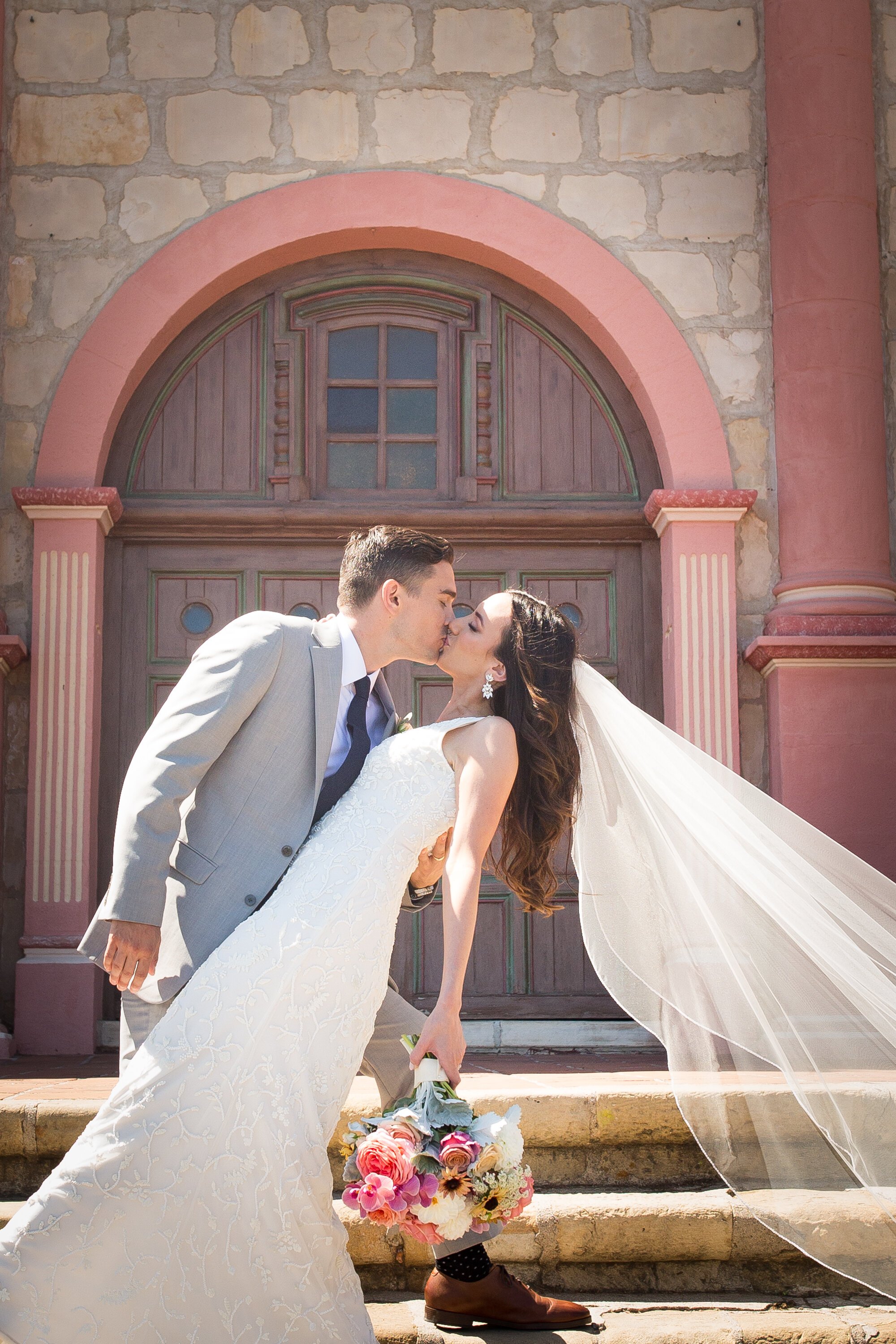 www.santabarbarawedding.com | Kelsey Crews Photo | Santa Barbara Mission | Ella &amp; Louie | Bride and Groom Kiss In Front of Colorful Church Building