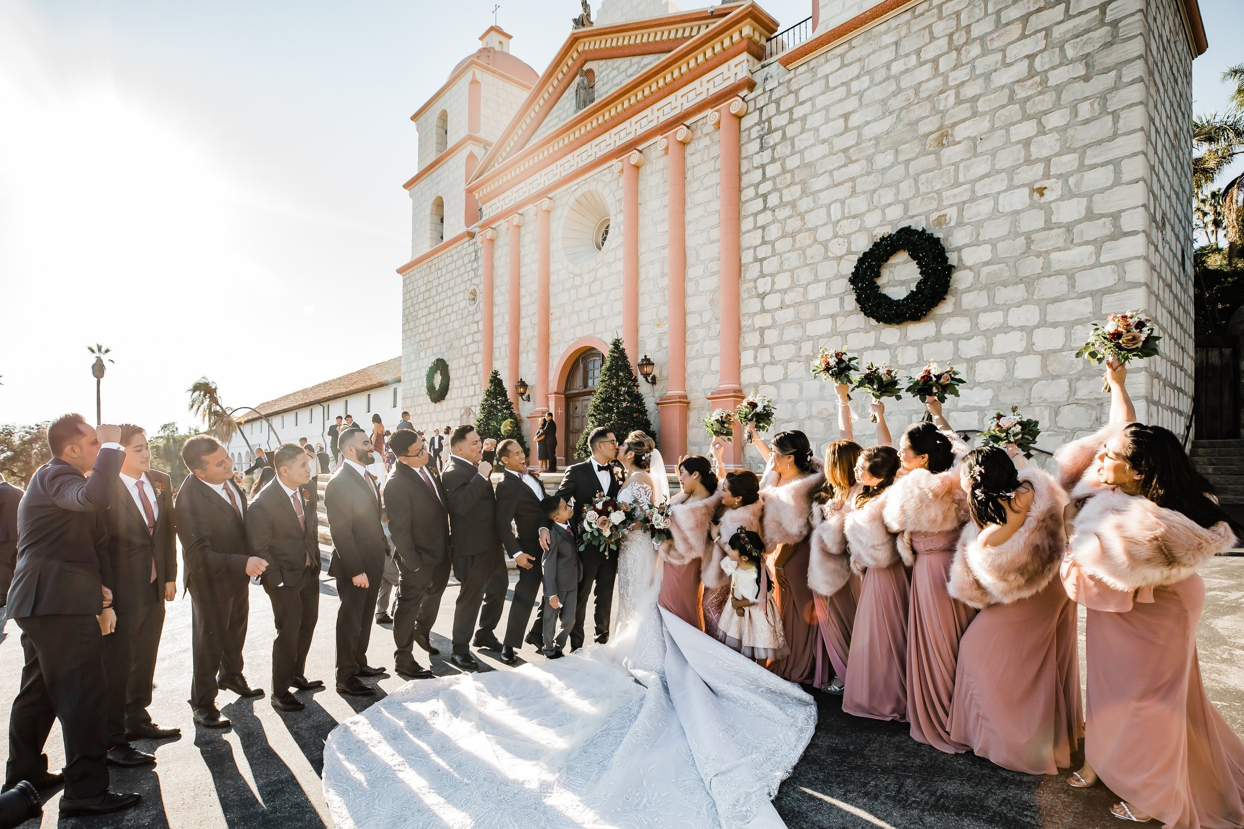 www.santabarbarawedding.com | Old Mission Santa Barbara | Michelle Ramirez Photography | Couple Kiss in Front of Church Surrounded by Bridal Party