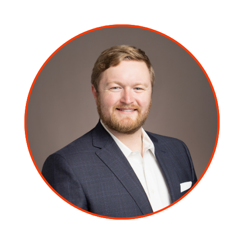 Portrait of a smiling man with light brown hair and beard, wearing a dark suit jacket and white shirt, against a gray background, framed with a red circle.